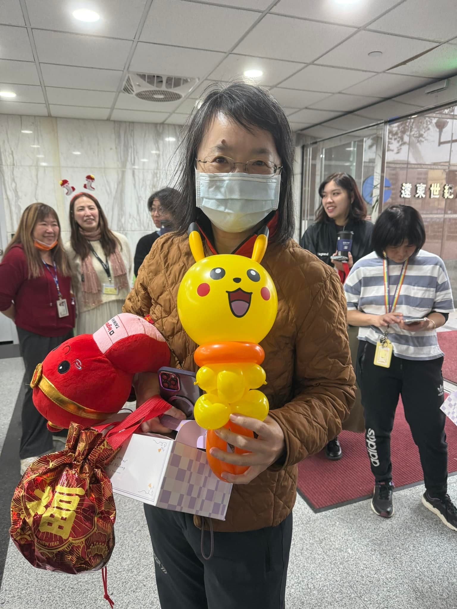 戴口罩的女士拿著皮卡丘造型氣球棒與紅色福袋，背景是排隊的同仁