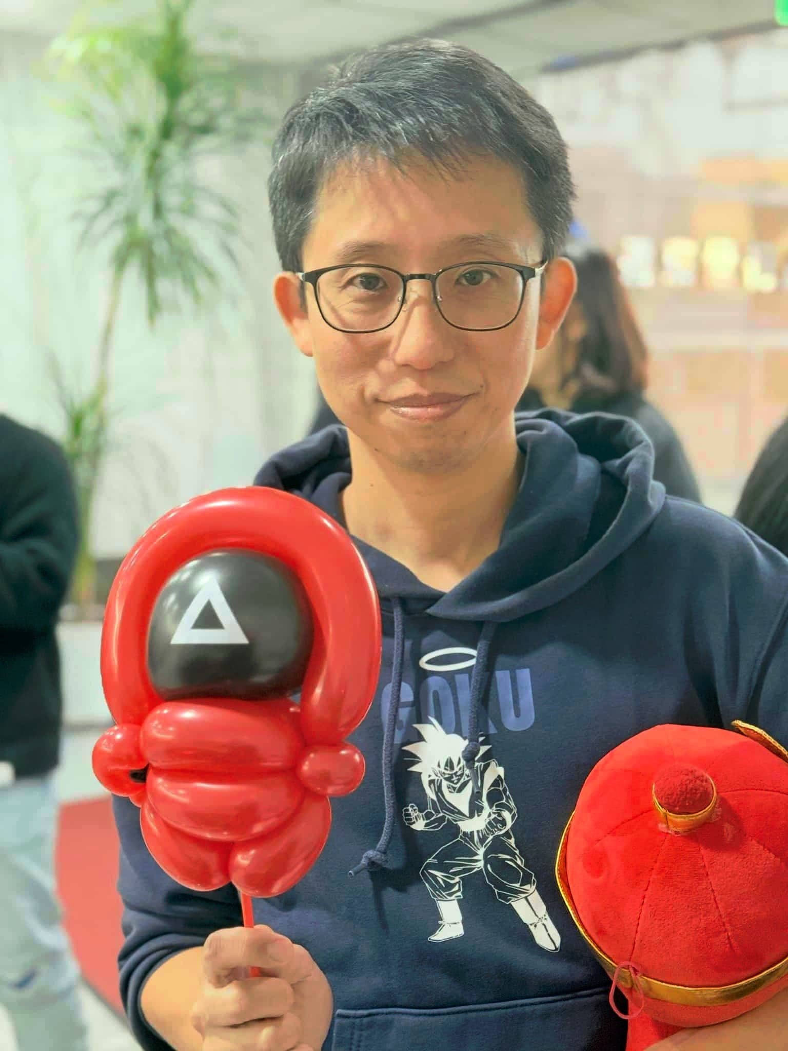 戴眼鏡的男士拿著紅色魷魚遊戲士兵造型氣球，黑色面罩上有白色三角形
