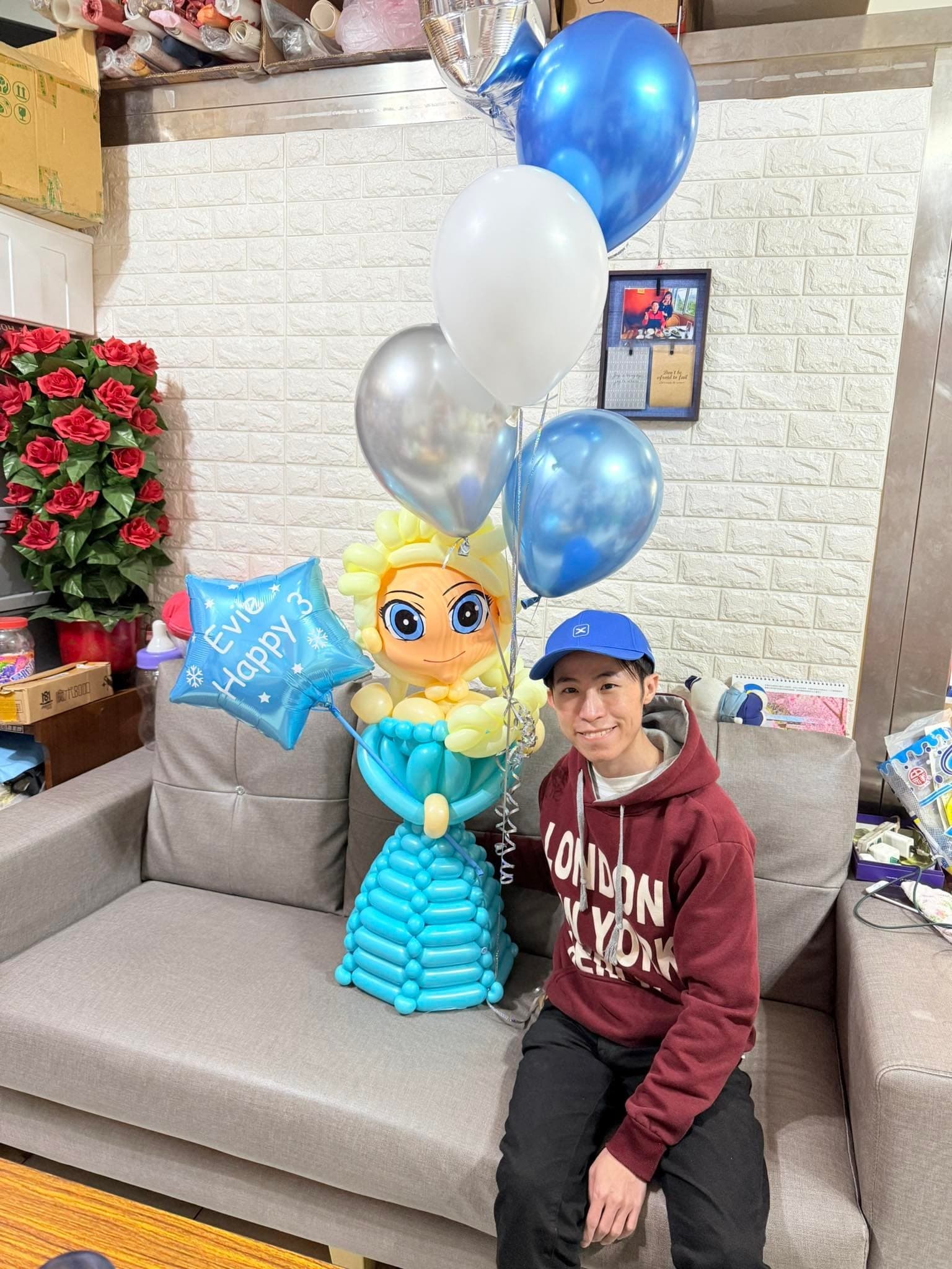 柯男戴藍色帽子坐在沙發上，旁邊是艾莎氣球人偶與藍銀白空飄氣球串