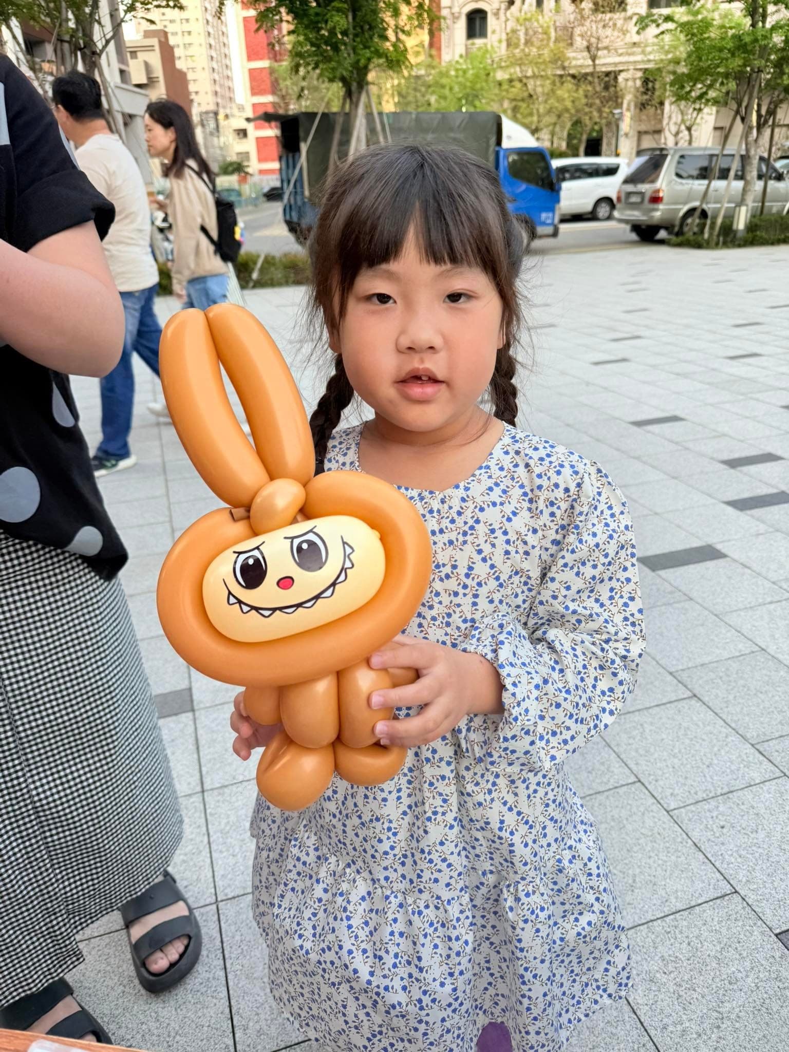 穿藍色碎花洋裝的女孩抱著橘咖啡色 Labubu 造型氣球站在市集廣場