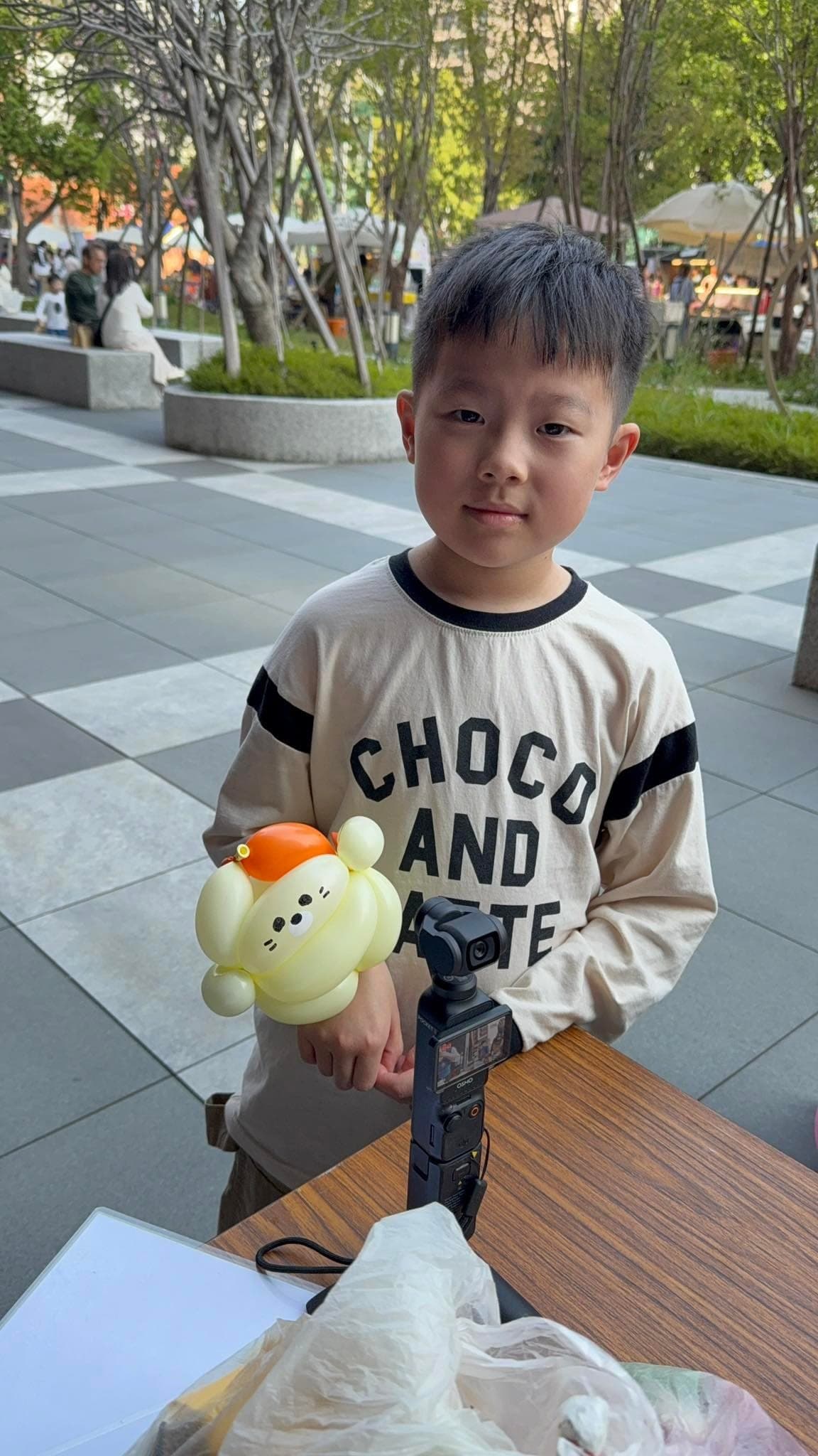 穿米色上衣的男孩在市集廣場拿著戴橘色帽子的黃色小狗造型氣球
