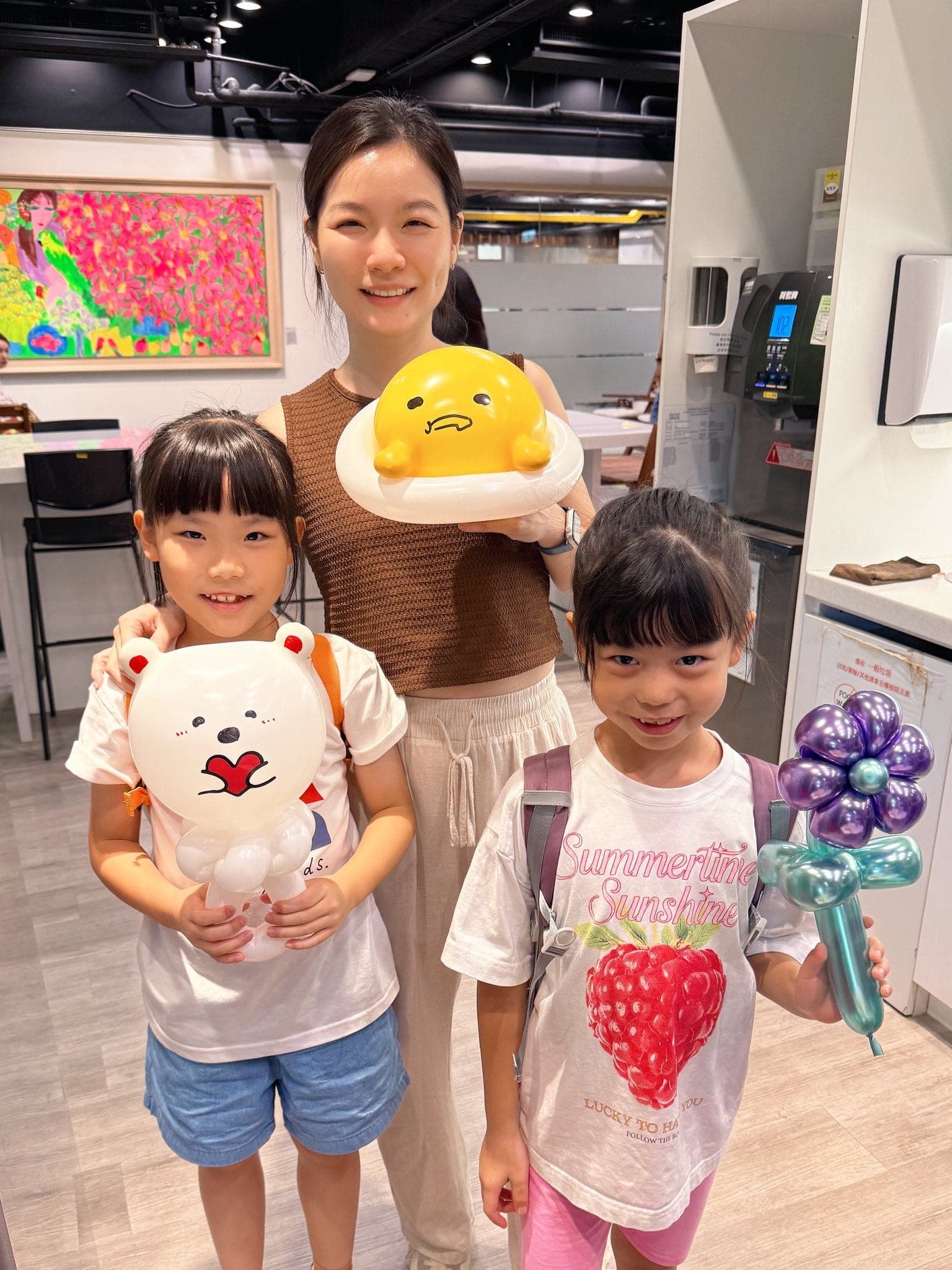 媽媽捧著黃色蛋黃哥造型氣球，兩個女兒拿著角落生物白熊抱愛心與紫色金屬花氣球