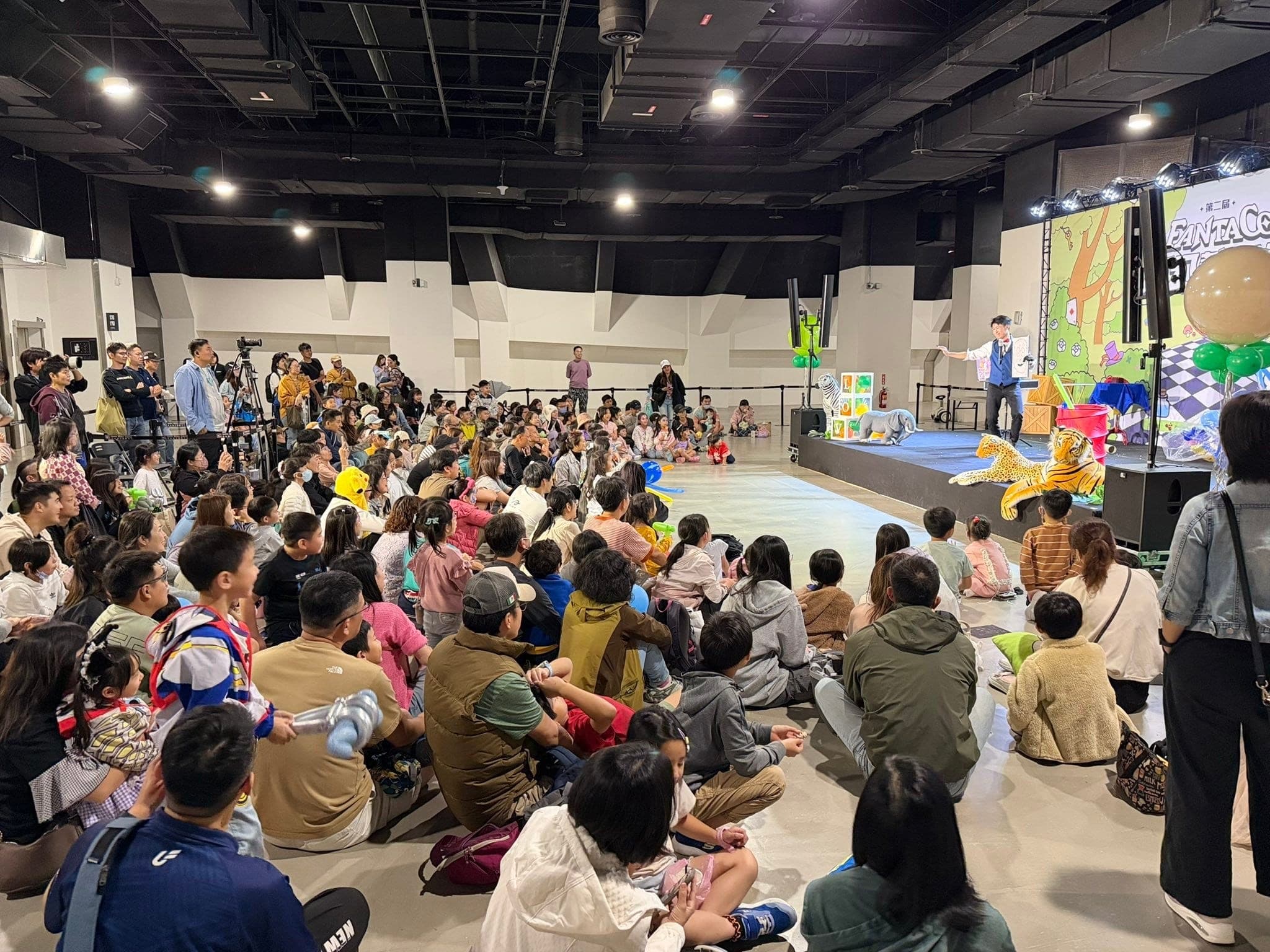 從場地後方看演出全景，大批觀眾坐在地上，柯男在遠處舞台表演，兩側有老虎造型布偶