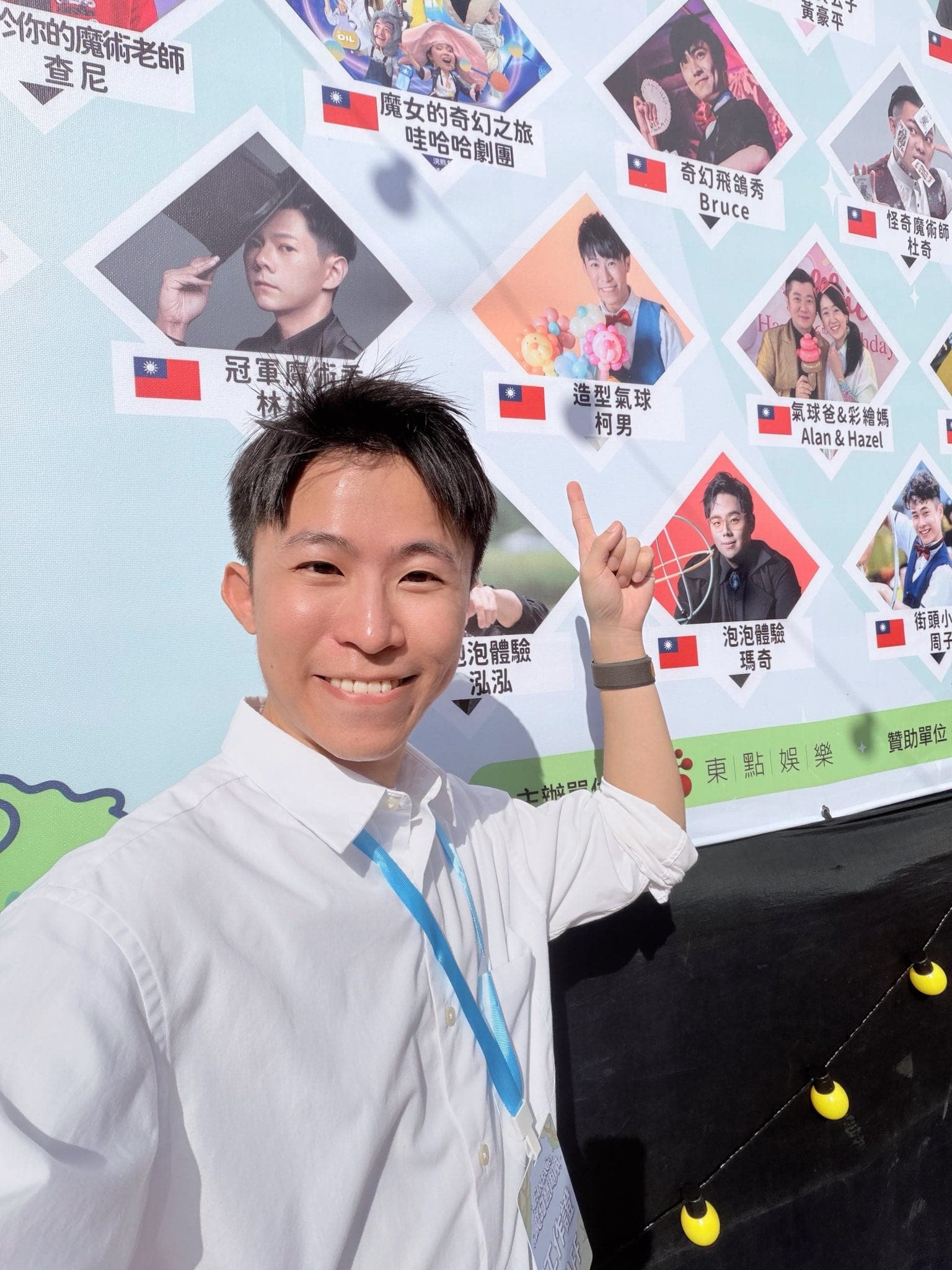 柯男指著藝術節表演者介紹牆上「造型氣球 柯男」的照片，牆上還有其他魔術師與劇團