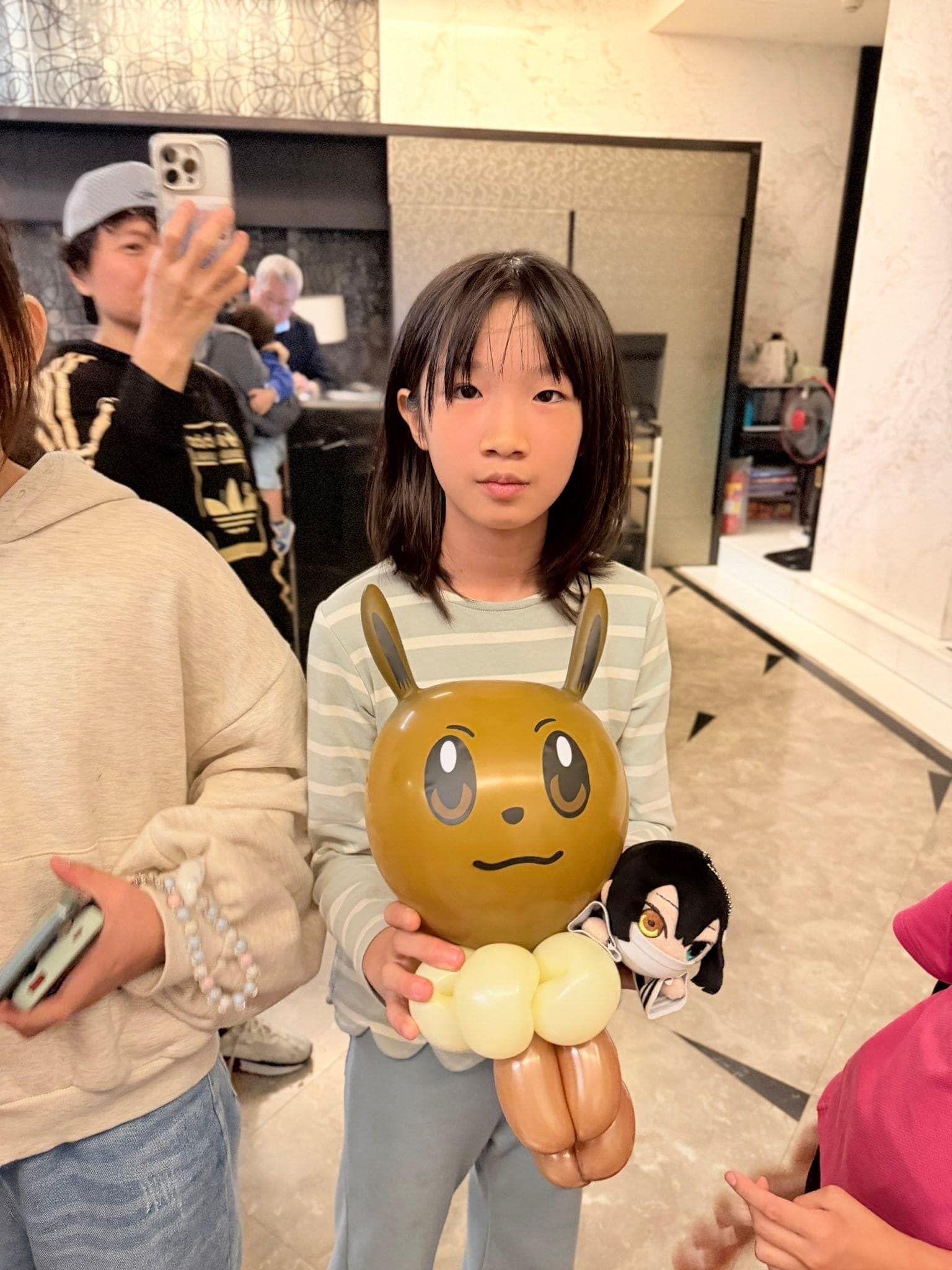 女孩捧著棕色長耳朵、大眼睛的動物造型氣球，旁邊夾著一個黑髮小娃娃吊飾