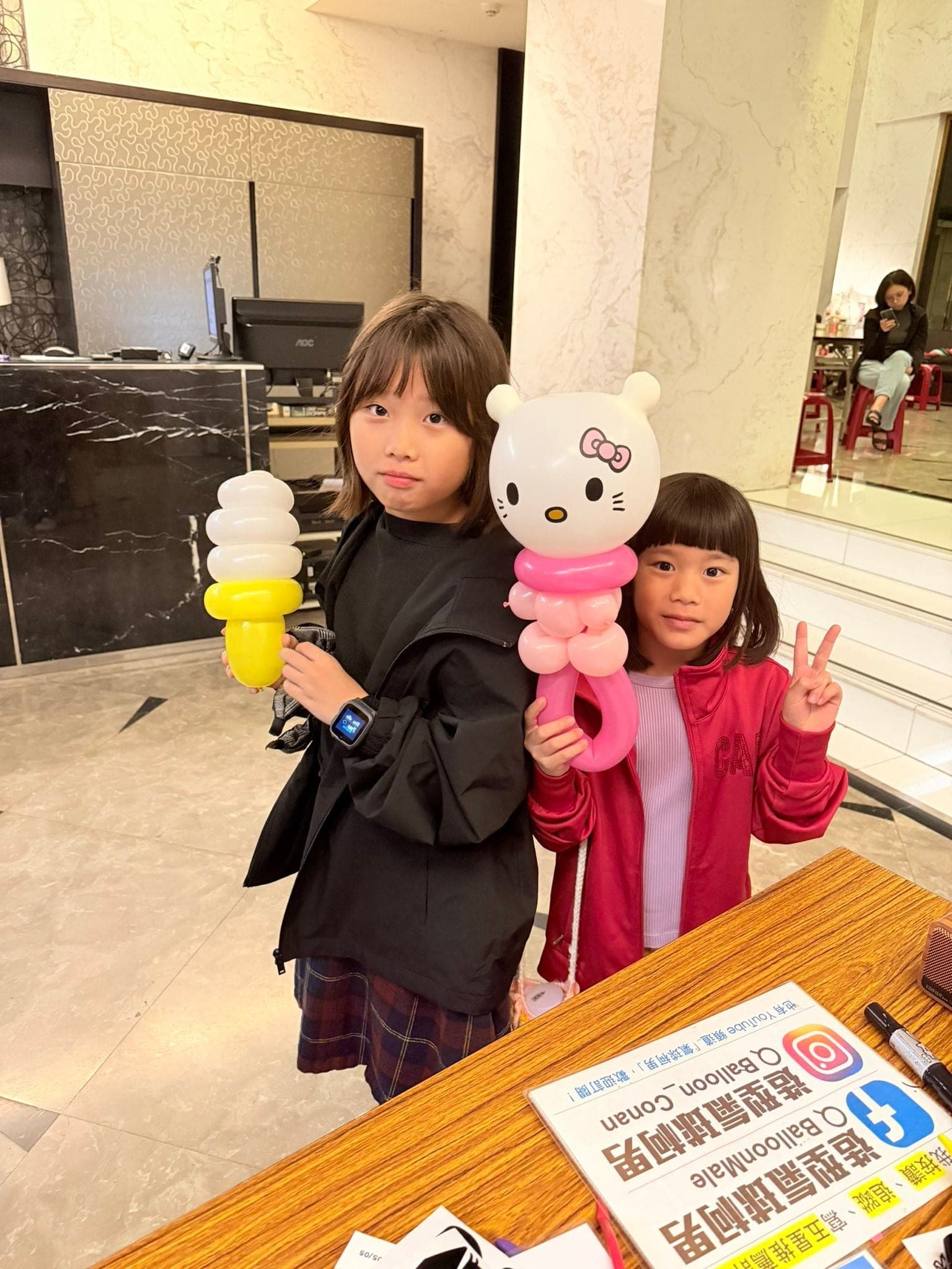 兩個女孩一個拿 Hello Kitty 造型氣球、一個拿黃白色冰淇淋造型氣球，桌上有柯男的粉絲頁立牌