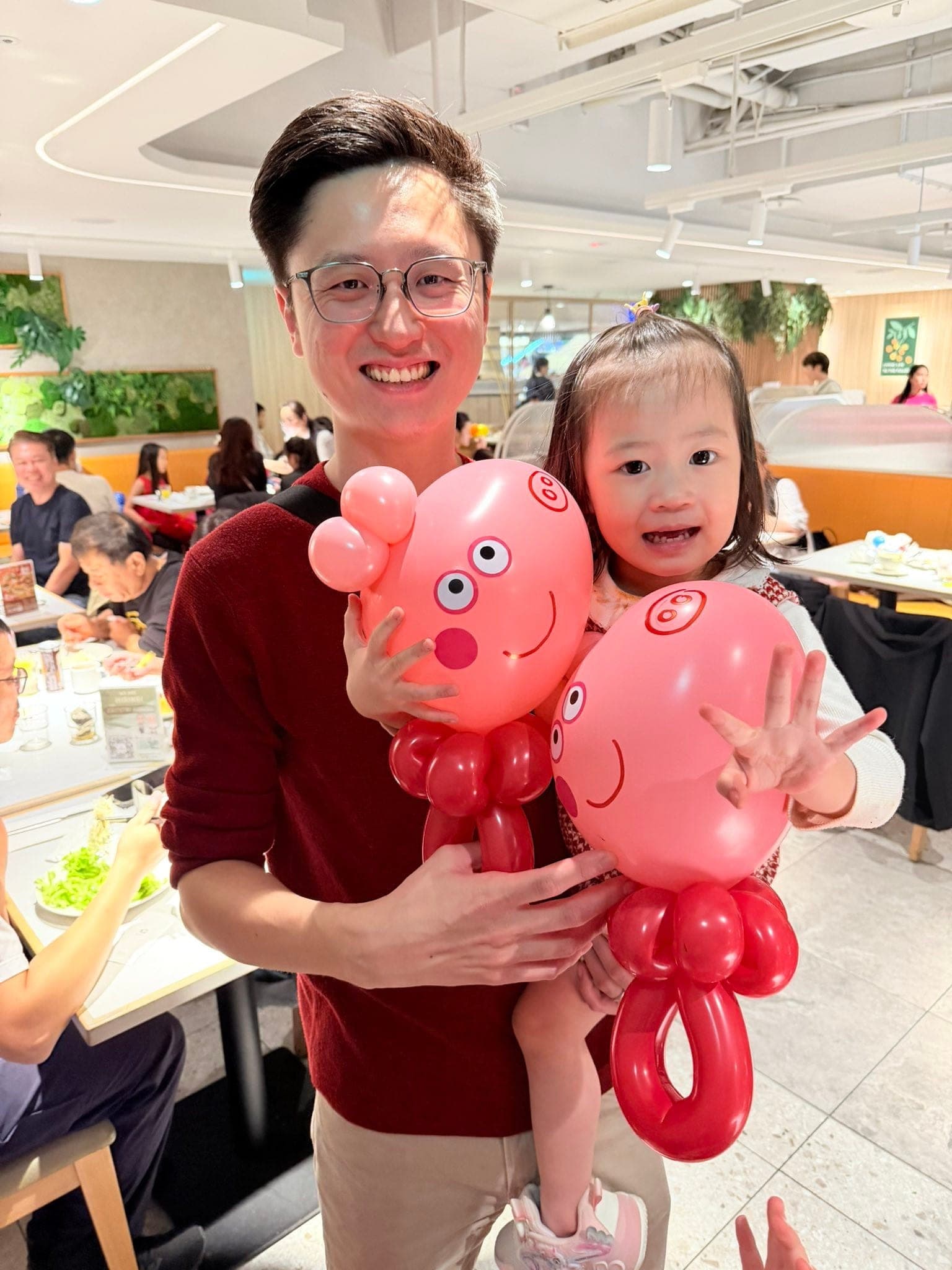 爸爸抱著小女兒，兩人各拿一隻粉紅色佩佩豬造型氣球，背景是自助餐廳座位