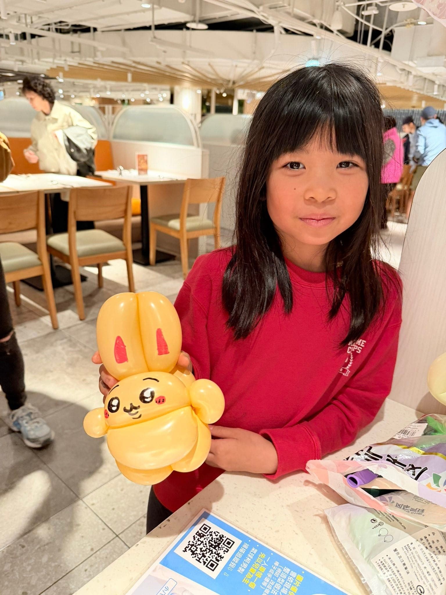 穿紅色上衣的女孩捧著黃色長耳兔子造型氣球，坐在餐廳桌邊