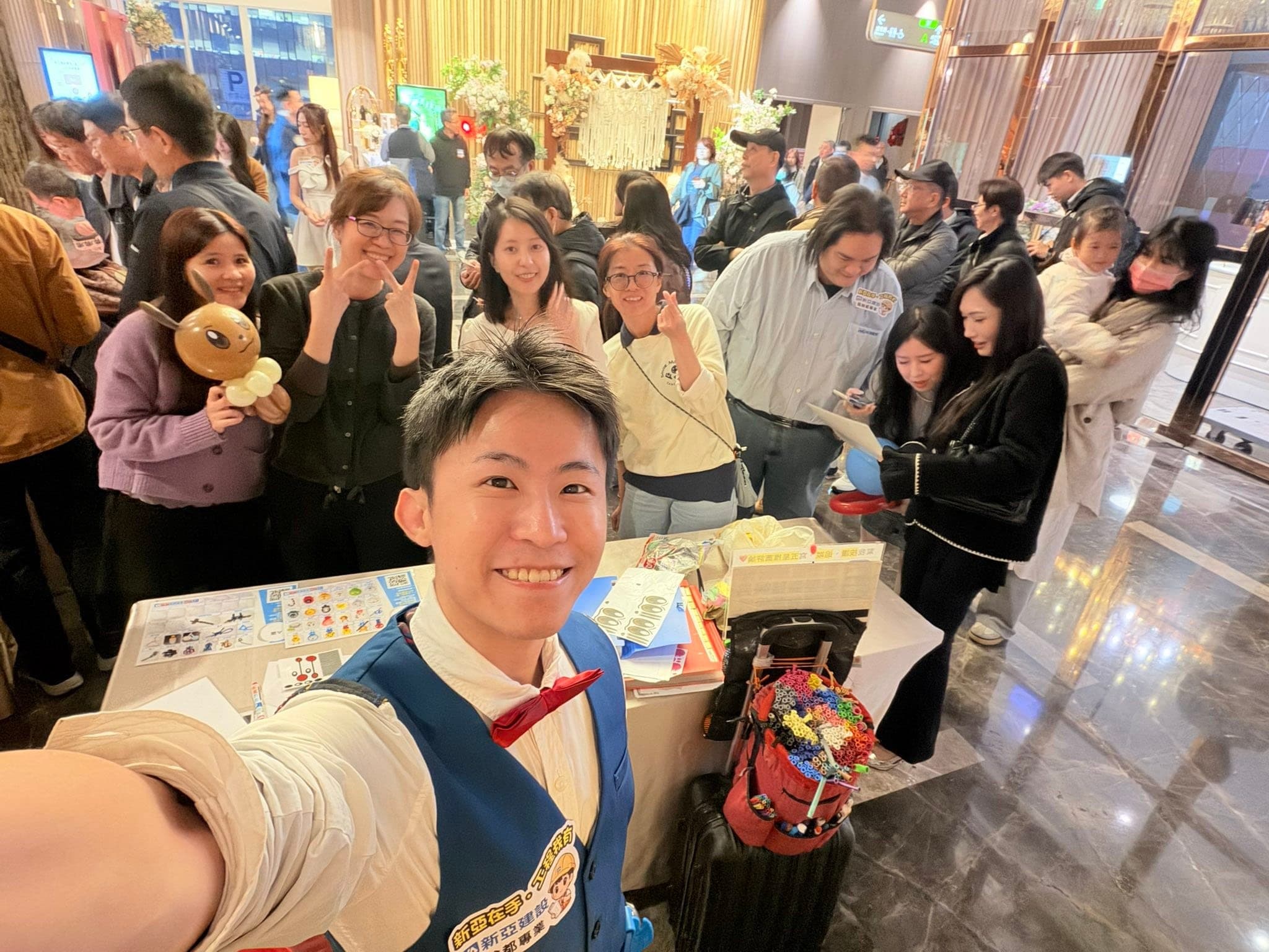 柯男在尾牙會場入口的折氣球攤位自拍，後方來賓排隊，一位女生拿著棕色造型氣球比 YA