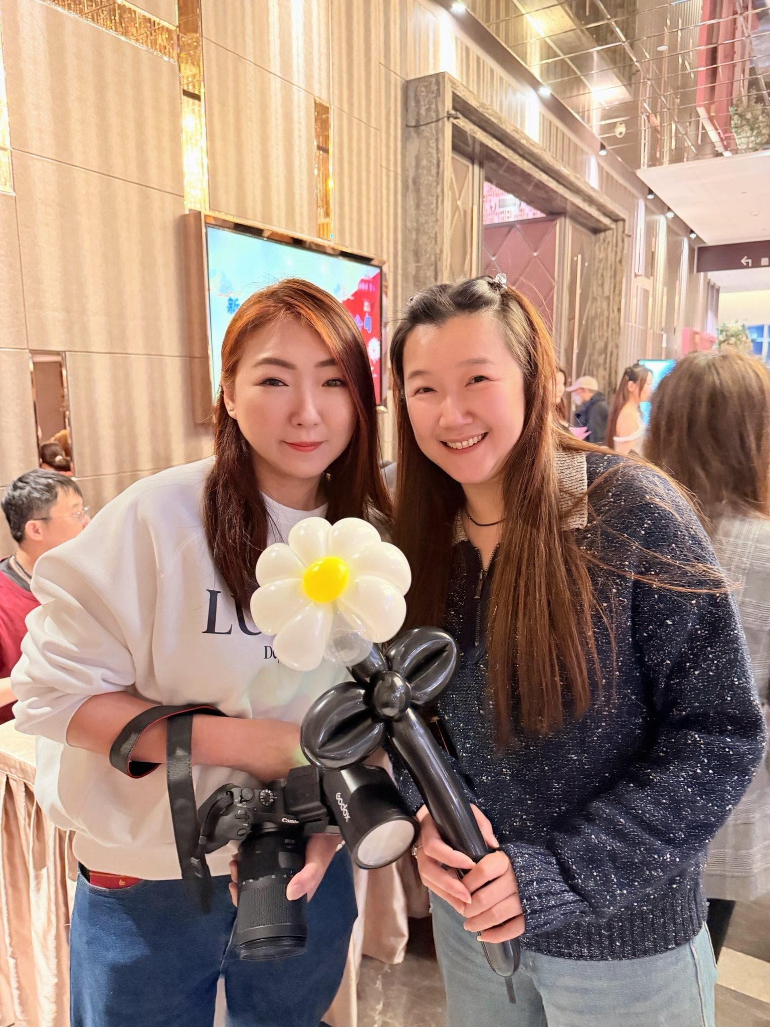 兩位女性來賓在宴會廳走道合照，其中一位拿著小雛菊造型氣球，另一位背著相機