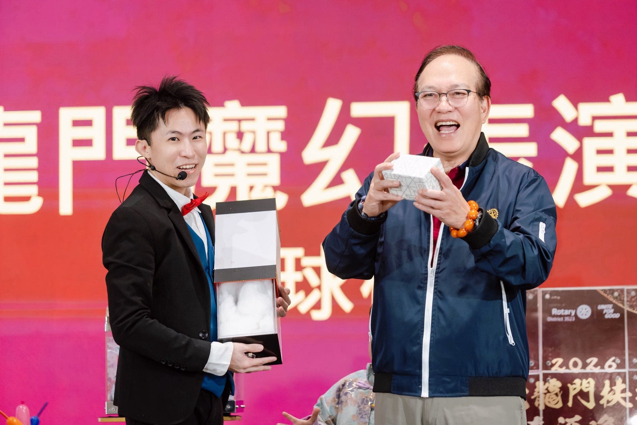 穿深藍外套的長輩在舞台上拿起白色小盒子笑得很開心，柯男手捧打開的禮盒站在一旁