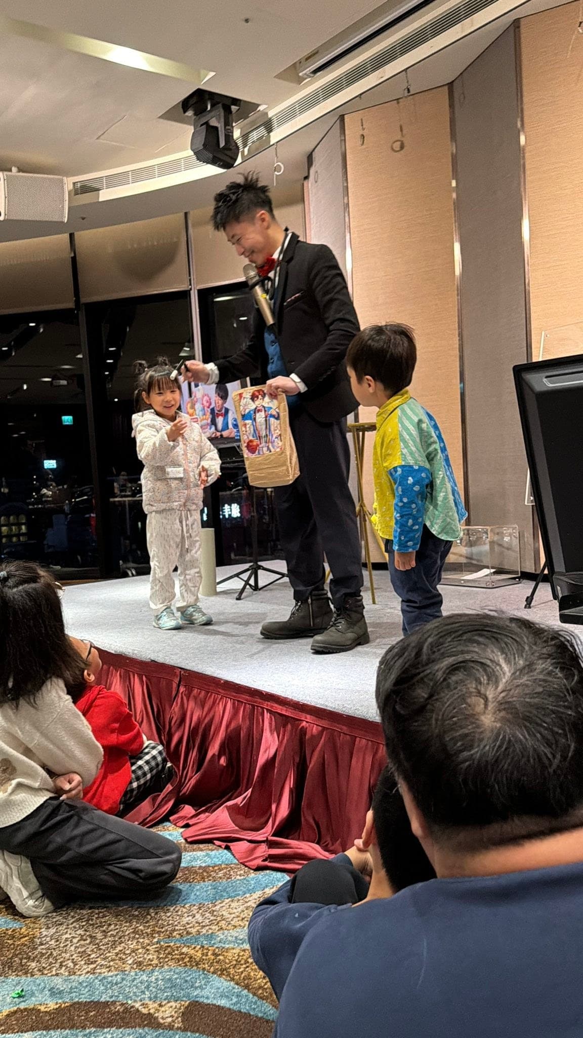 宜蘭礁溪尾牙舞台上，柯男拿著魔術道具紙袋，與穿白色連身衣的小女孩和穿彩色外套的男孩互動