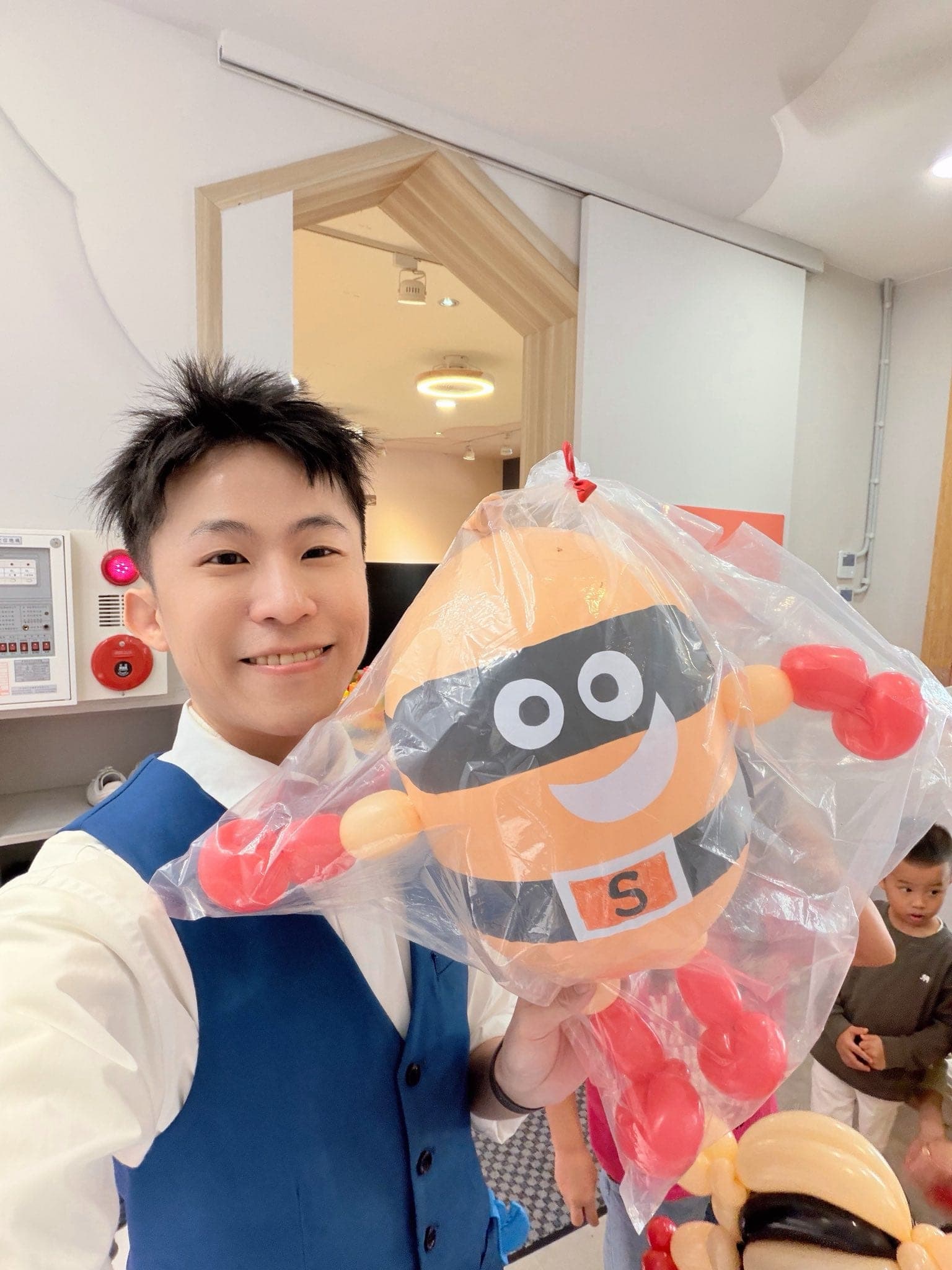 柯男在活動現場自拍，手上拿著裝進透明袋的土豆超人造型氣球，後方有小朋友