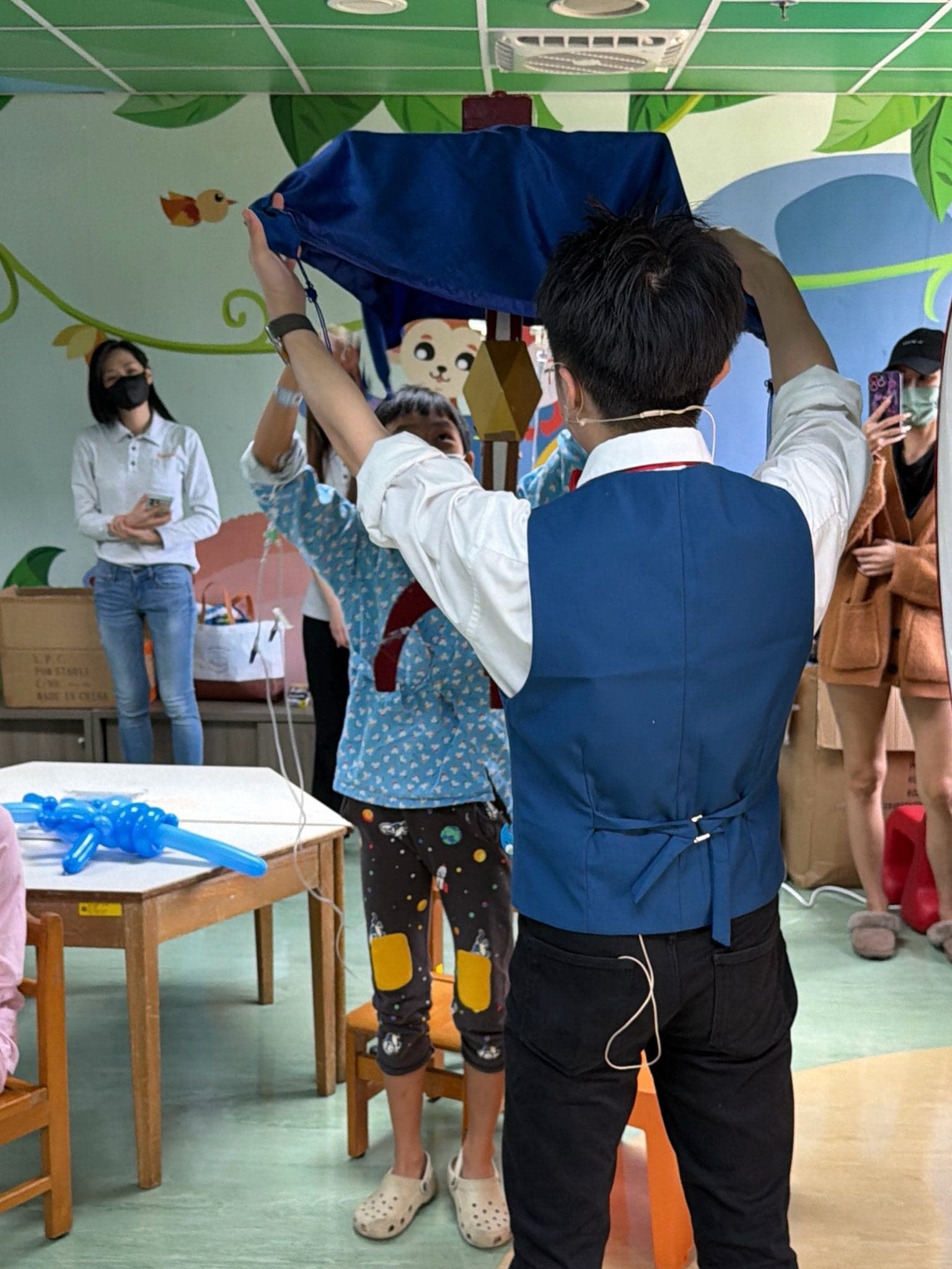 小朋友上台和柯男一起拉起藍色桌巾進行魔術體驗，旁邊桌上放著藍色造型氣球