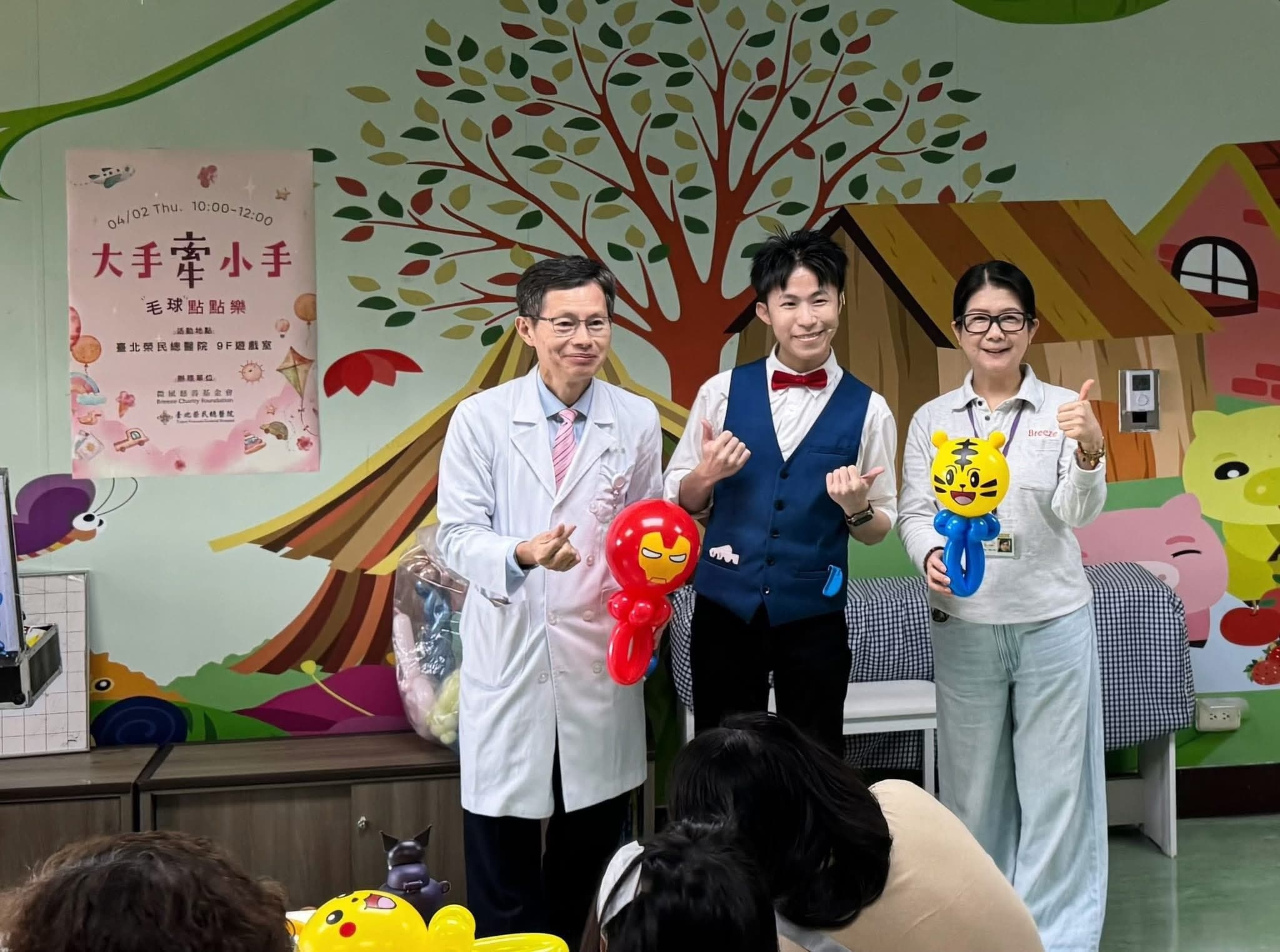 台北榮總兒童病房遊戲室，柯男與穿白袍的醫師、工作人員合照，兩人分別拿著鋼鐵人與巧虎造型氣球