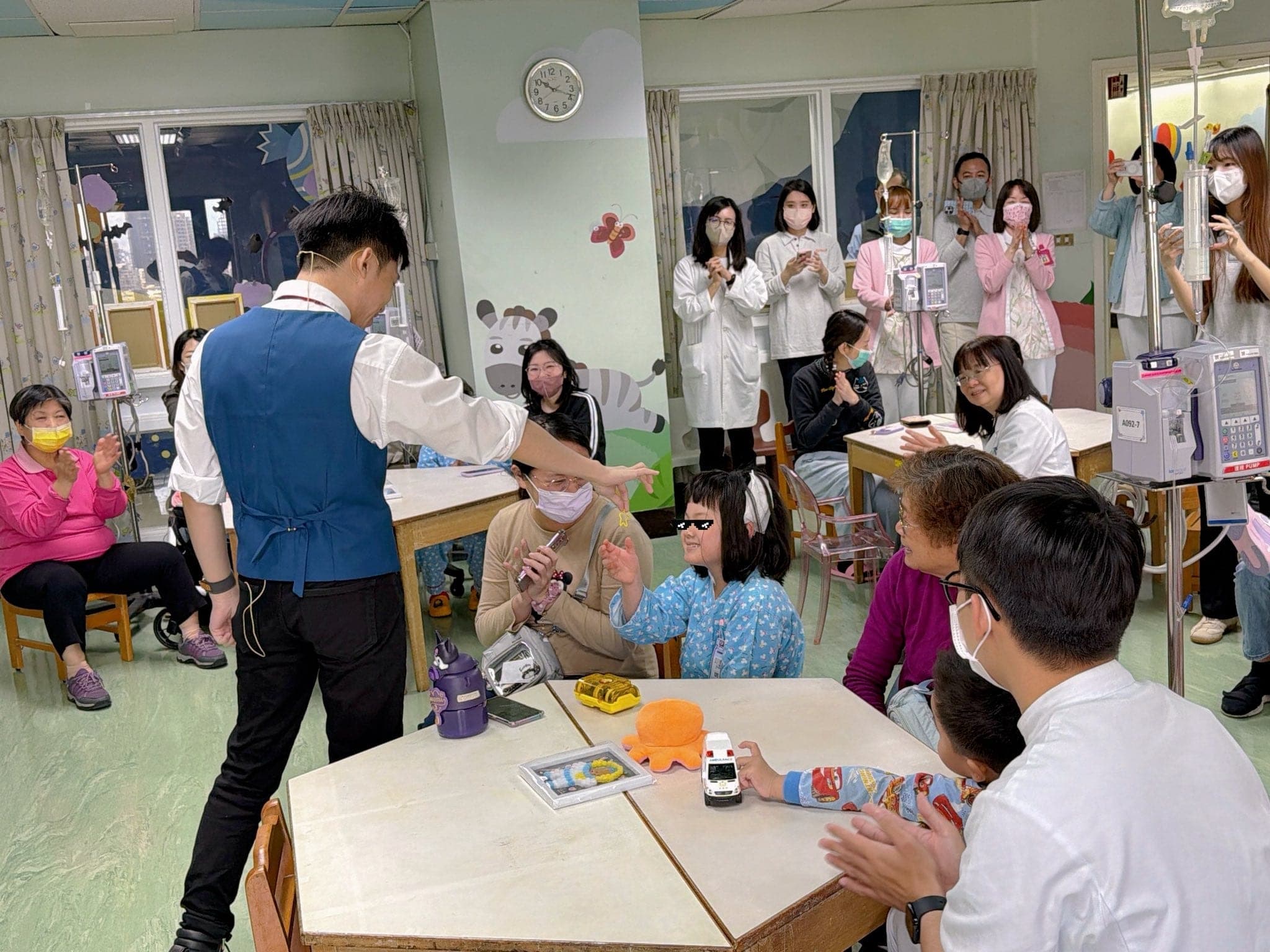 柯男穿藍色背心在病房遊戲室表演，桌邊的孩子和家長看著他的手勢笑開懷