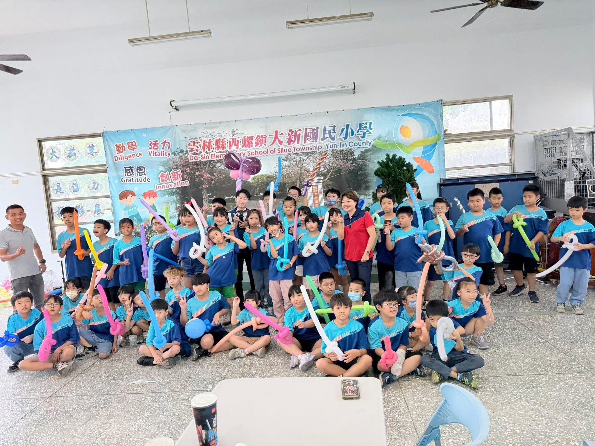 雲林西螺大新國小學生穿藍色制服，人手一把彩色氣球寶劍在校名布條前大合照