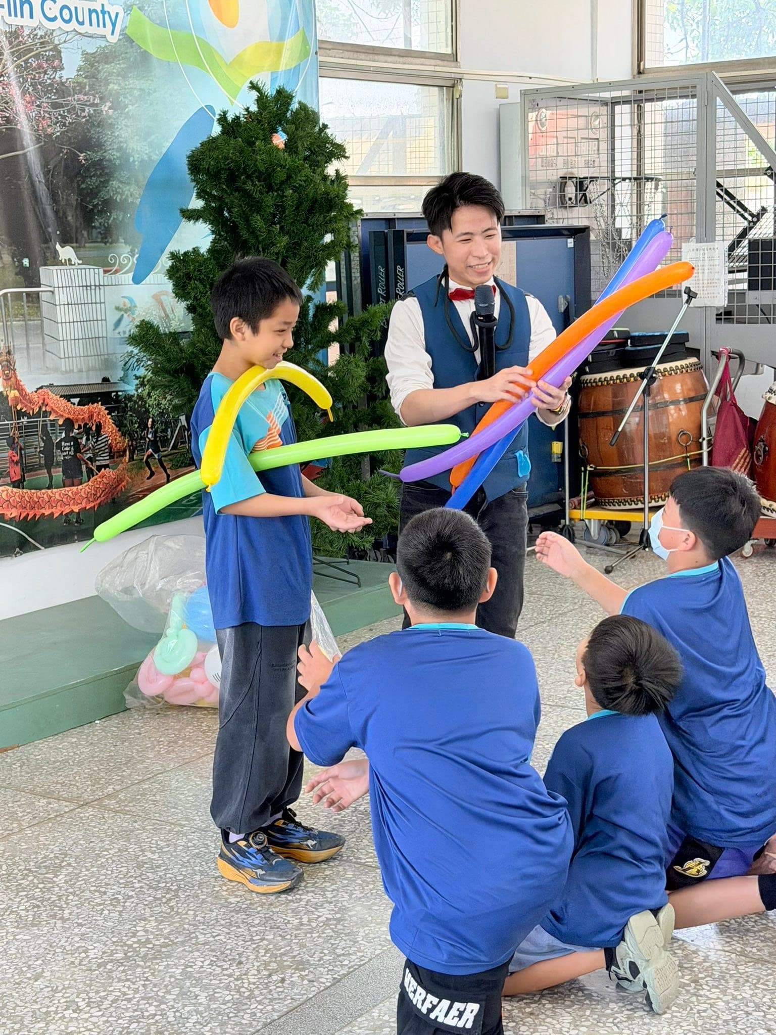 戴頭戴式麥克風的柯男把橘紫色長條氣球遞給上台的男孩，旁邊同學蹲坐觀看