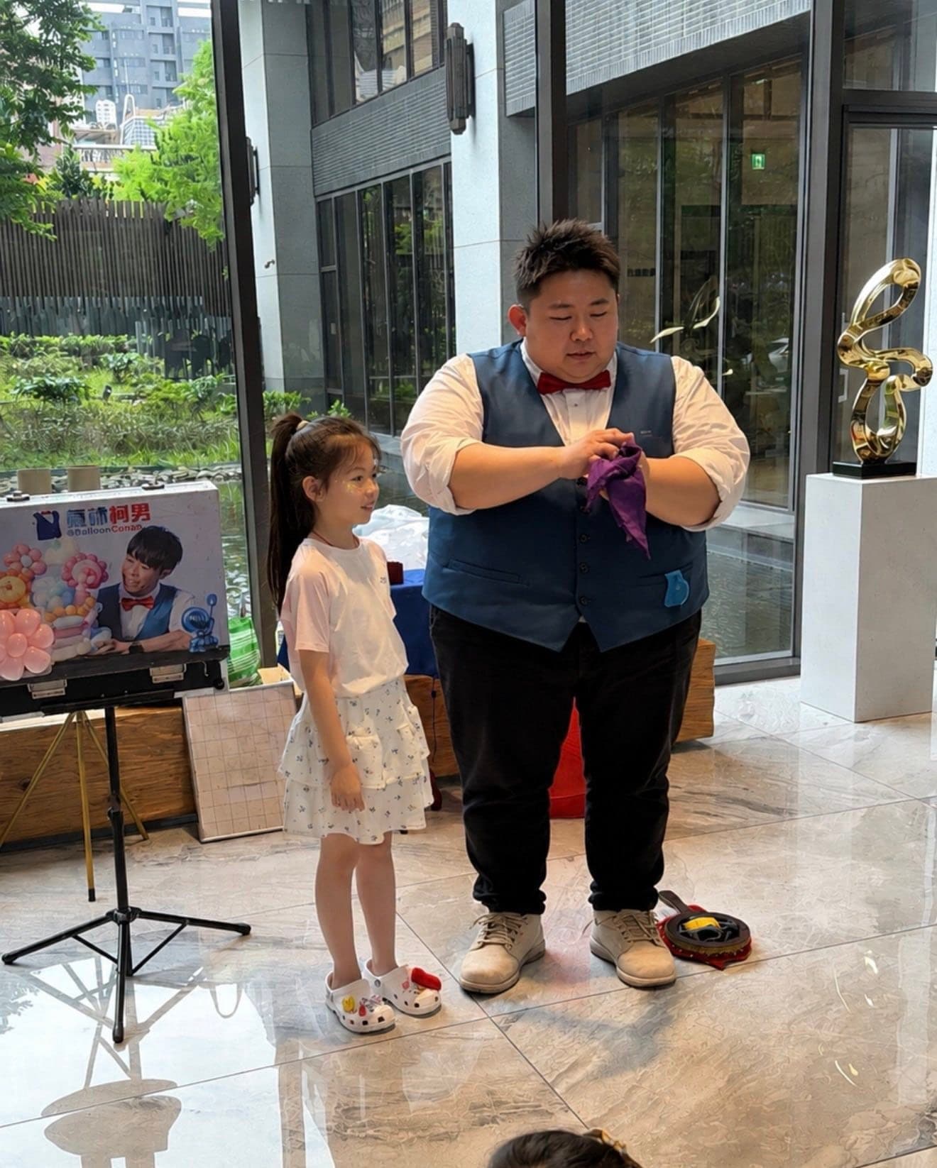 穿藍色背心的魔術師手拿紫色絲巾，身旁穿白色洋裝的小女孩上台協助表演