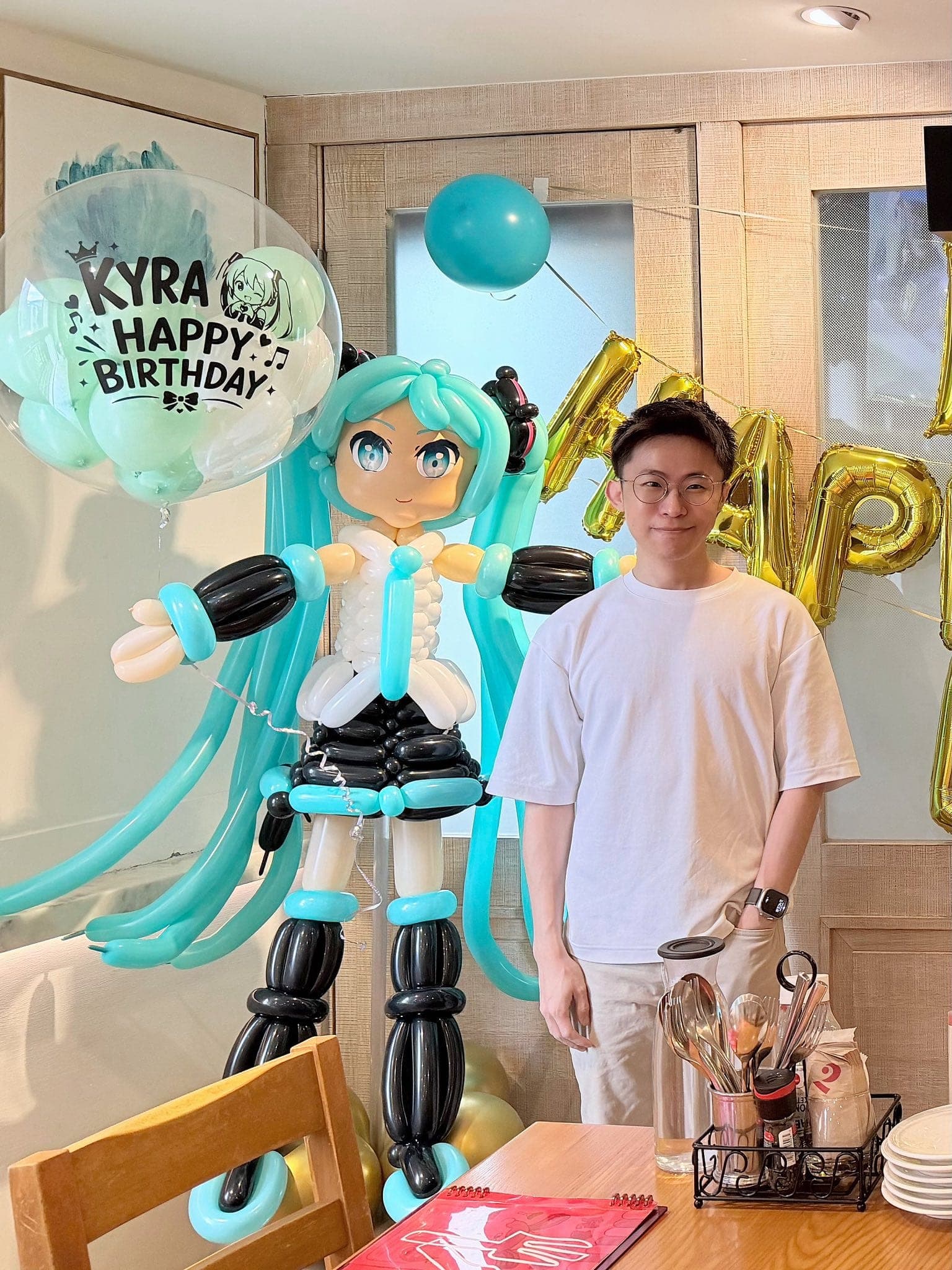 戴眼鏡穿白色 T 恤的男生站在初音未來氣球人偶旁合照，後方是金色字母氣球