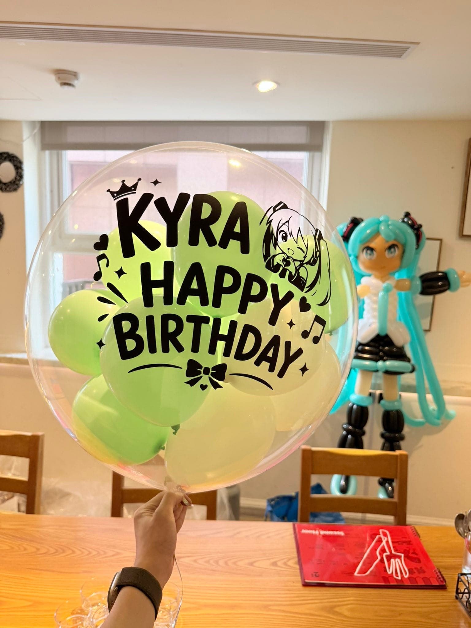 初音未來中型氣球人偶站在金銀色氣球底座上，藍綠色雙馬尾、黑白服裝，旁邊是透明客製波波球