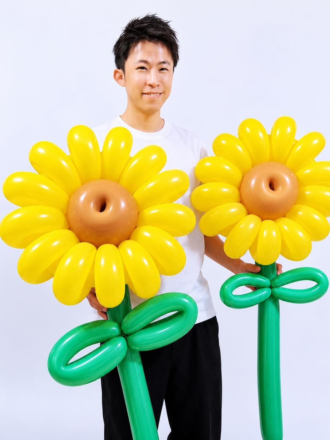 柯男雙手各拿一朵巨無霸太陽花氣球，黃色花瓣、咖啡色花心，綠色花莖綁著蝴蝶結葉子