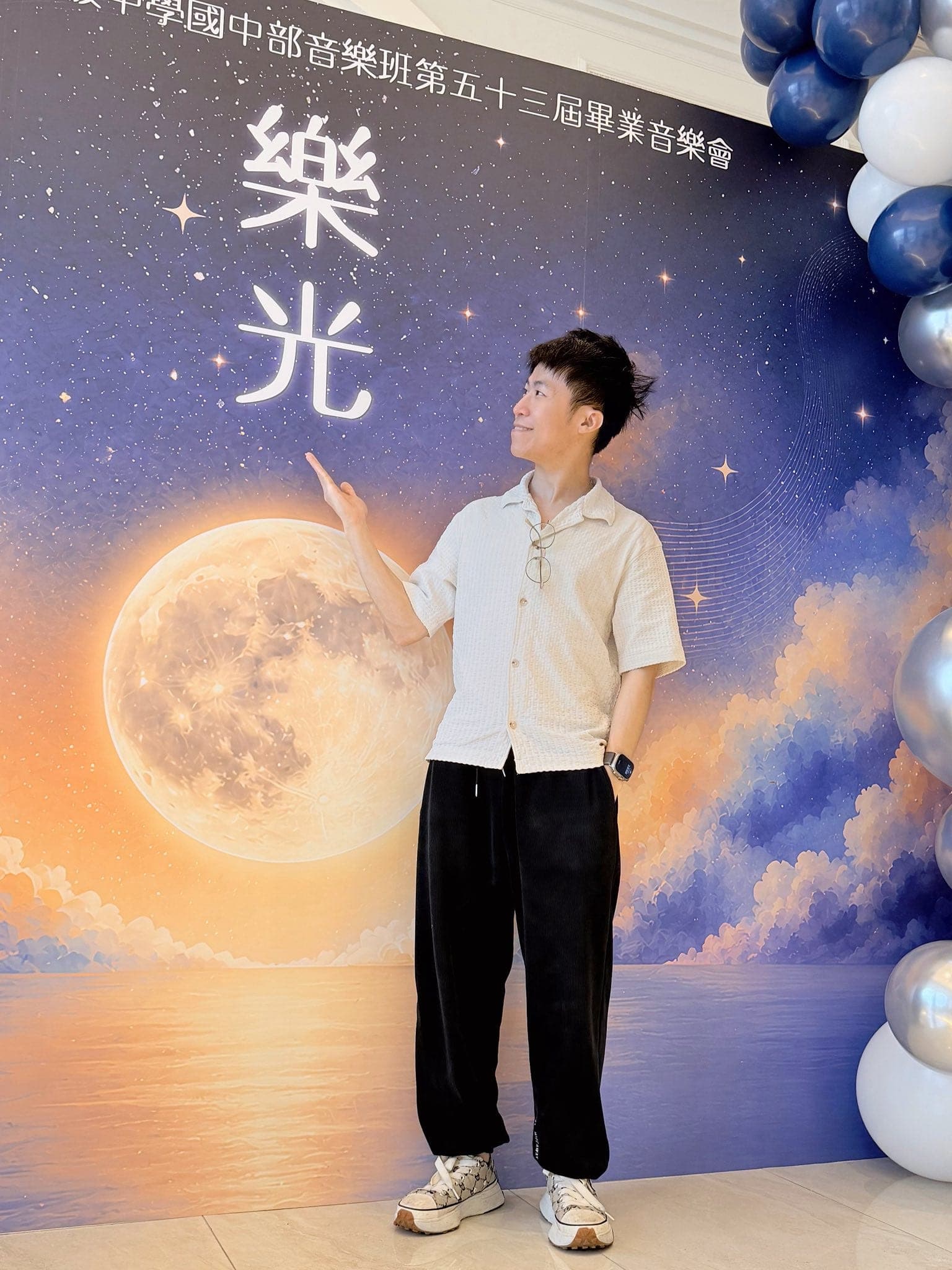 柯男站在夜空滿月背板前抬手指向畫面上方，右側是深藍與銀色氣球