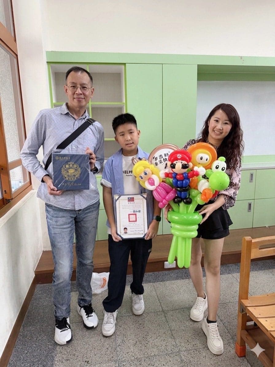 國小畢業生拿著獎狀站在教室裡，身旁大人捧著瑪利歐氣球花束、另一位大人拿著畢業紀念冊