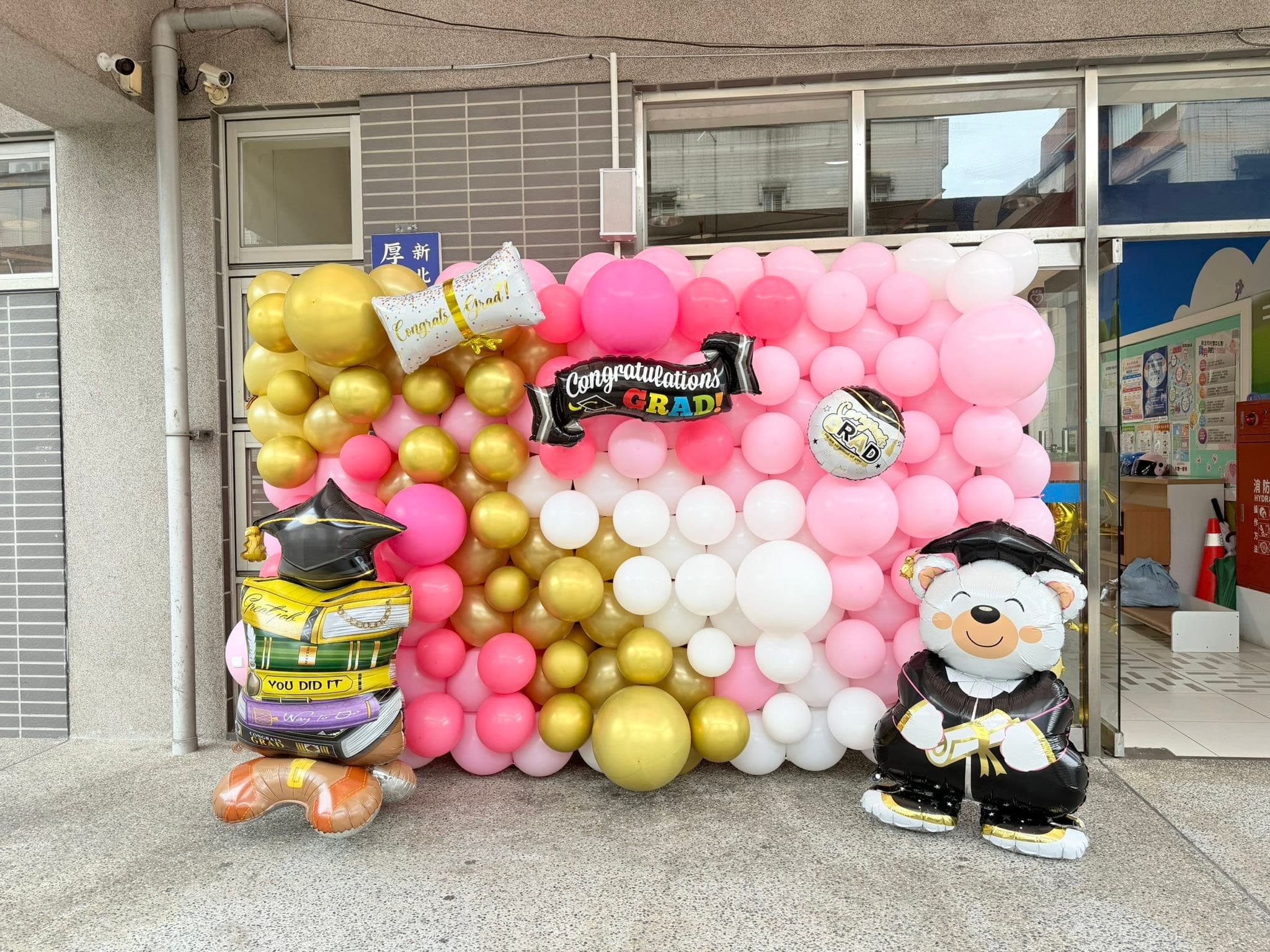 校舍門口的粉白金色氣球牆，掛著 Congratulations GRAD 鋁箔字條，旁邊有穿畢業袍的熊與書本造型氣球