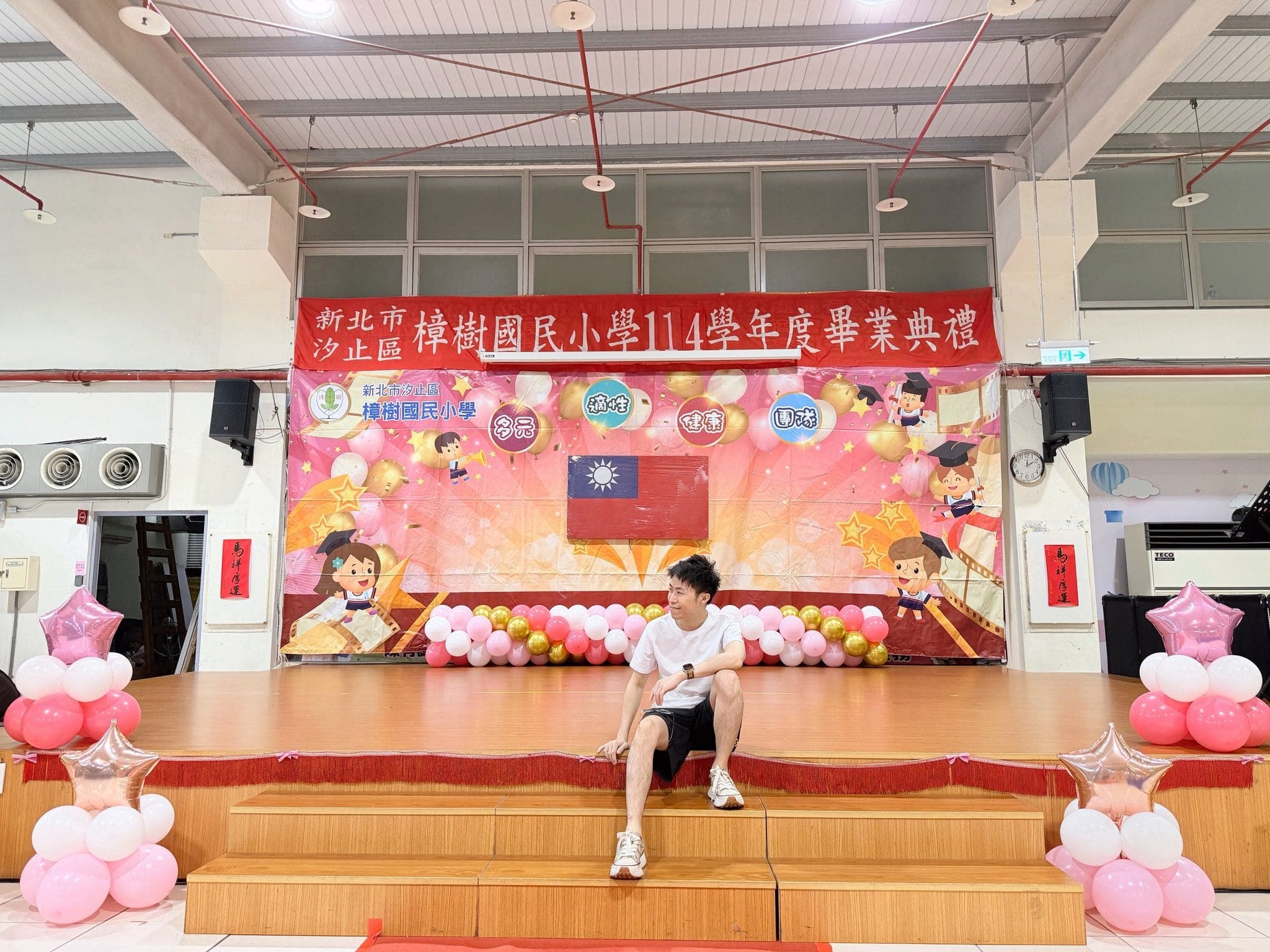 畢業典禮舞台全景，舞台邊緣有粉紅白金氣球串，台下兩側擺放粉紅星星鋁箔氣球柱