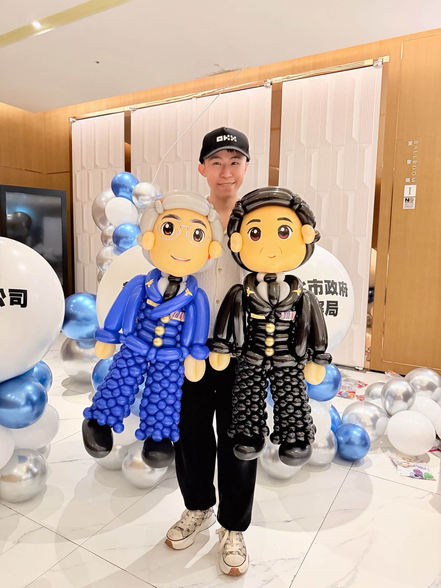 戴黑色棒球帽的柯男站在兩尊警察局長人像氣球中間，後方是藍銀白氣球與印字大氣球