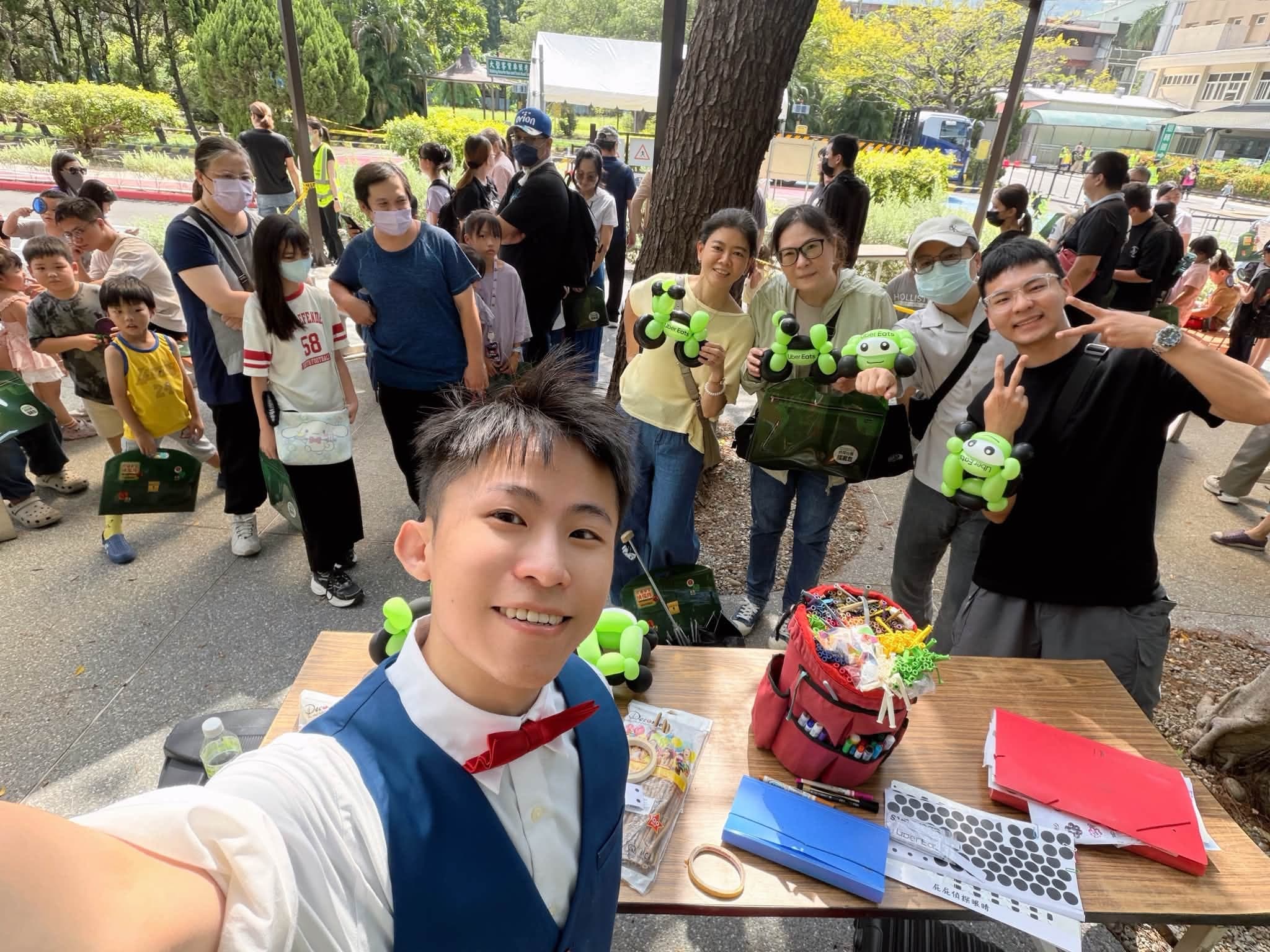 柯男在樹下攤位自拍，後方家長與孩子排隊，大人們手上拿著綠色 Uber Eats 機車造型氣球