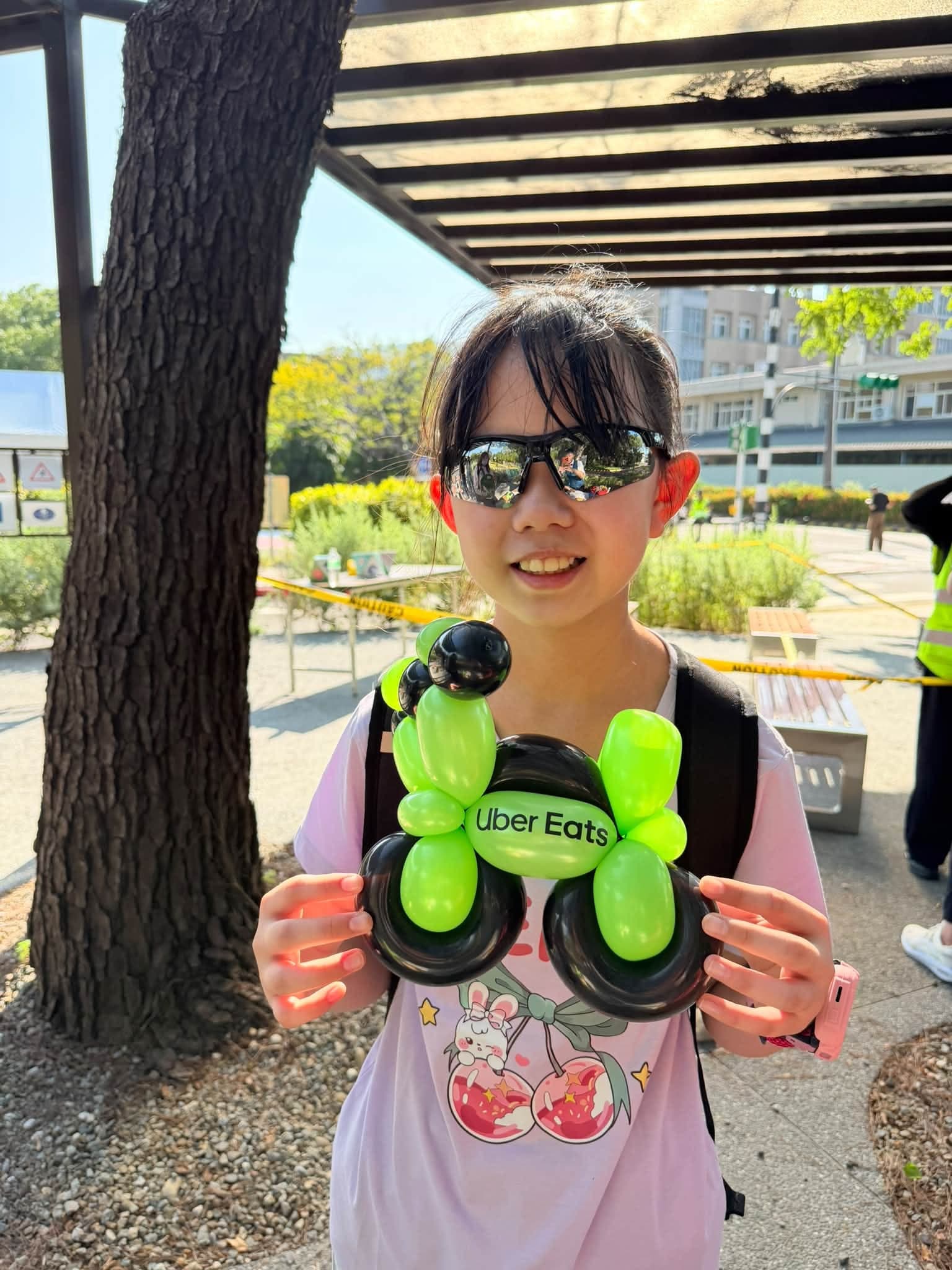 戴墨鏡的女孩雙手拿著綠色車身、黑色輪子的 Uber Eats 機車造型氣球