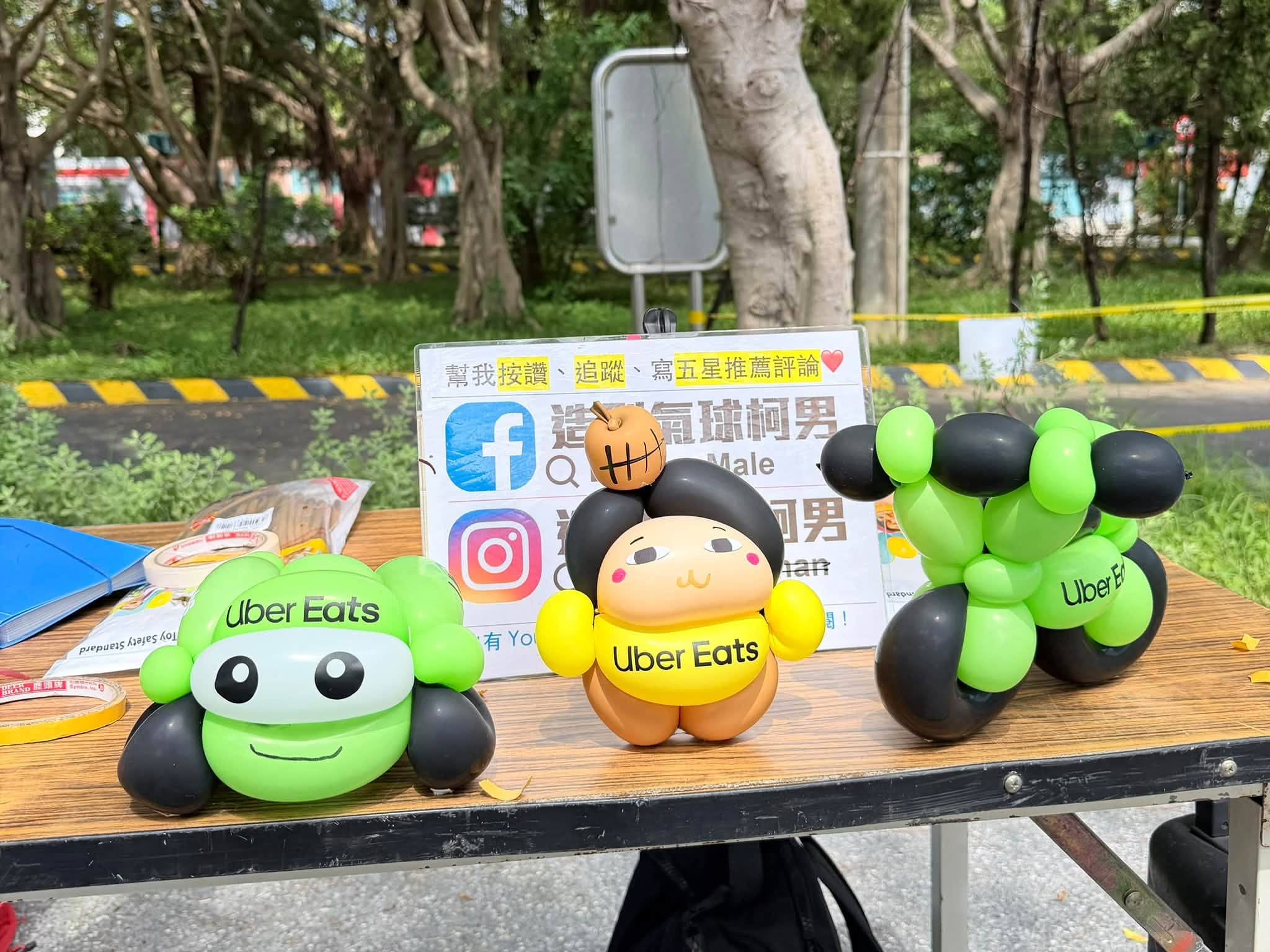 新北市監理所戶外攤位桌上擺著三款 Uber Eats 造型氣球：綠色大眼小車、黃色圍兜娃娃與綠黑機車，都貼有品牌貼紙