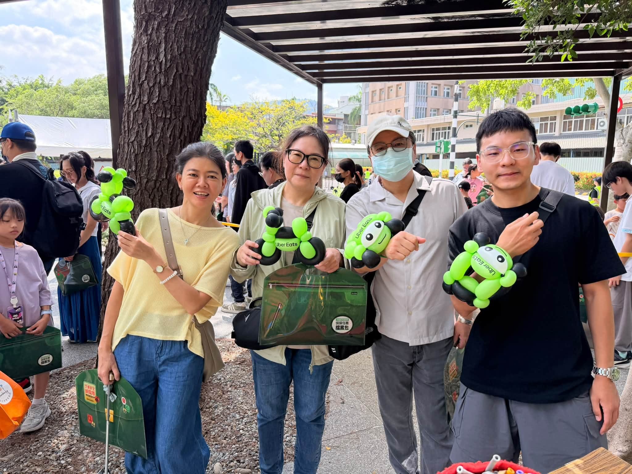 四位大人在樹下各拿一個綠色 Uber Eats 機車造型氣球合照，背景是排隊人潮