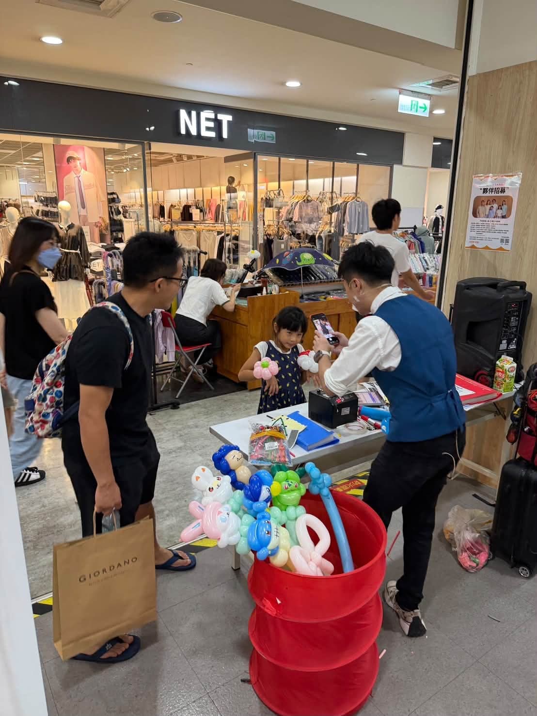 商場走道上的折氣球攤位，柯男為小女孩折氣球，紅色收納桶插滿造型氣球，旁邊家長提著佐丹奴購物袋