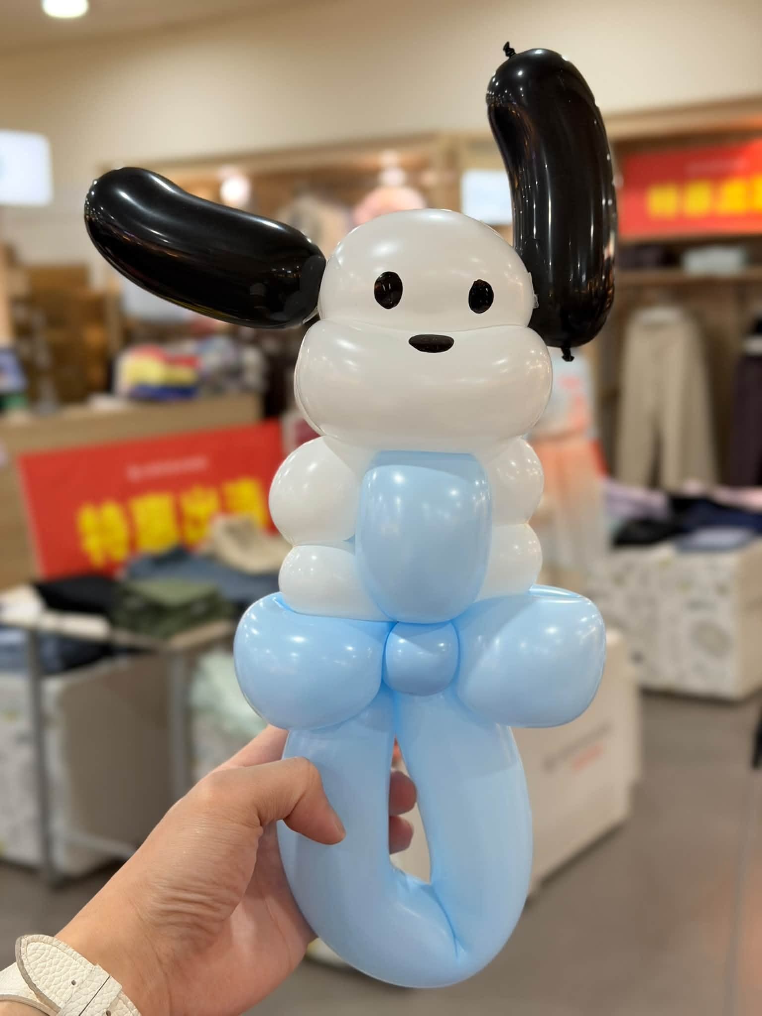 手持三麗鷗帕恰狗造型氣球（白色身體、黑色長耳朵），下方是淺藍色可套手的底座