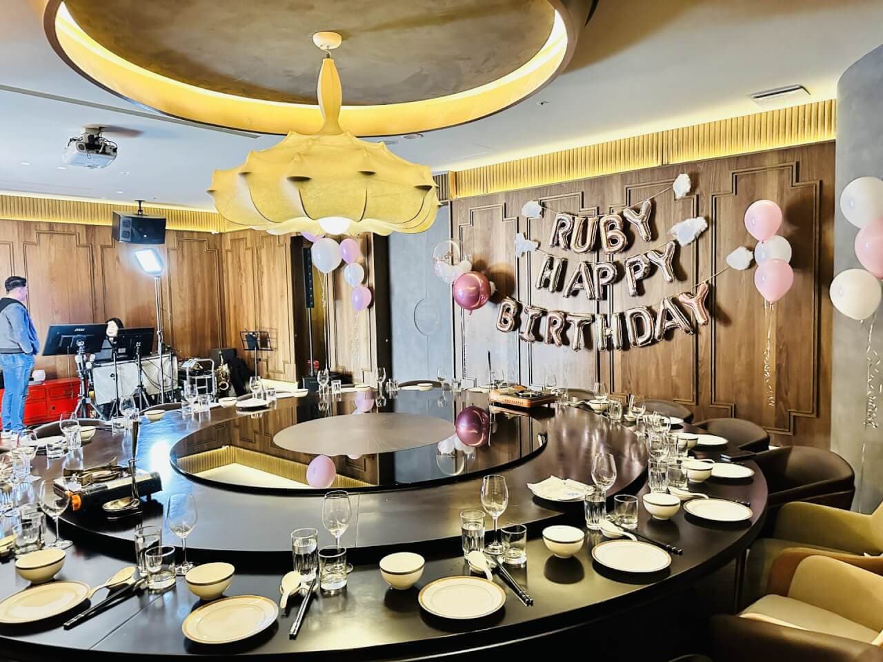 餐廳大圓桌包廂，木紋牆上掛銀色 RUBY HAPPY BIRTHDAY 字母氣球與粉白空飄氣球