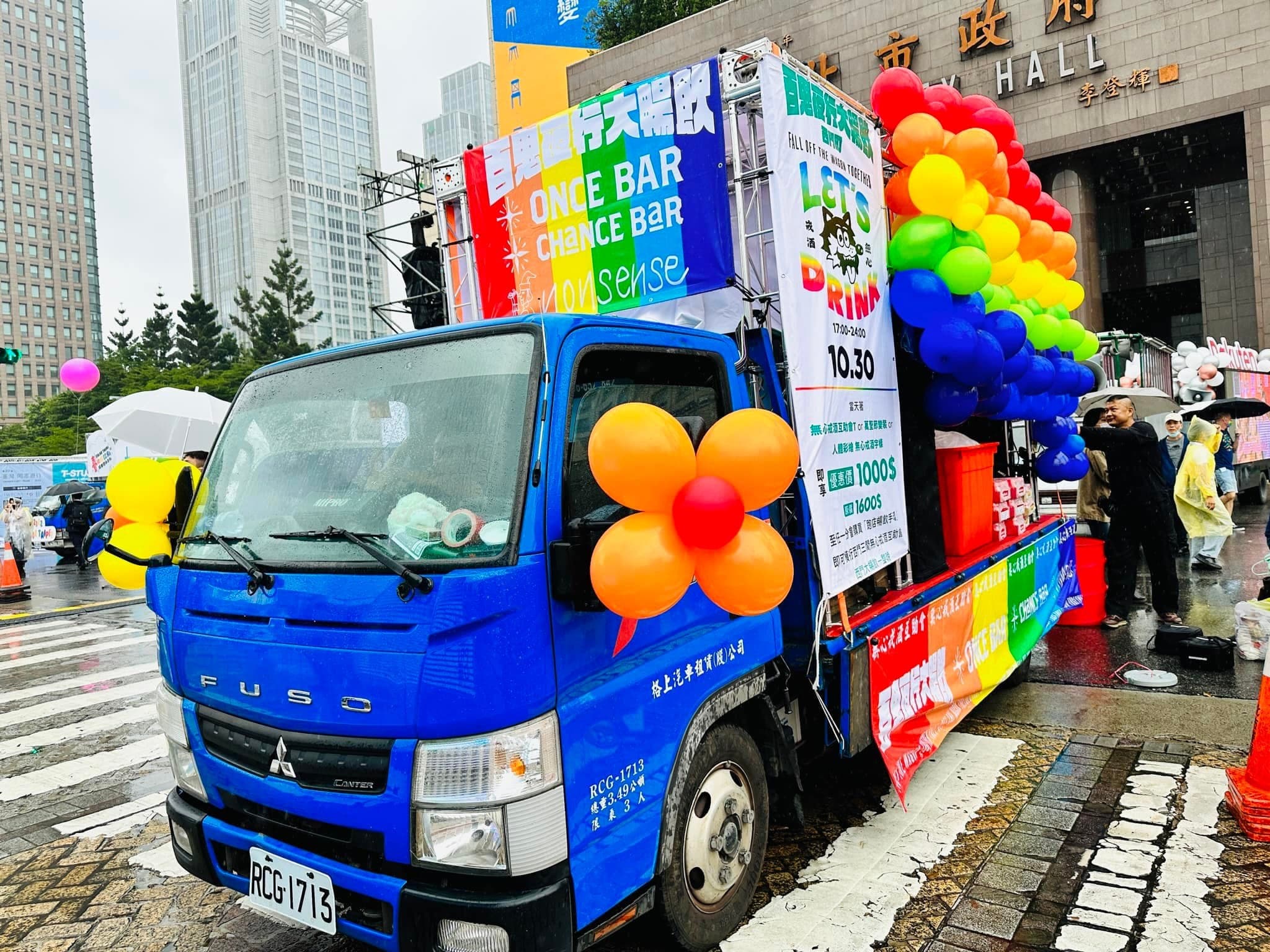 雨中的藍色舞台車停在市政府前,車頭有橘紅花朵氣球,車尾是彩虹氣球柱