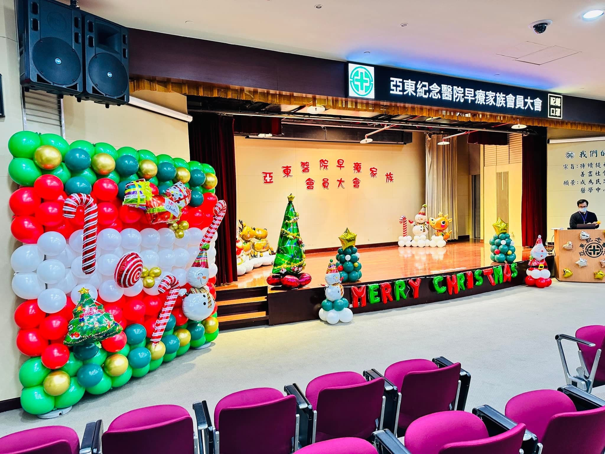 禮堂舞台左側是紅白綠聖誕氣球牆,台前有聖誕樹、雪人氣球柱和 MERRY CHRISTMAS 字樣