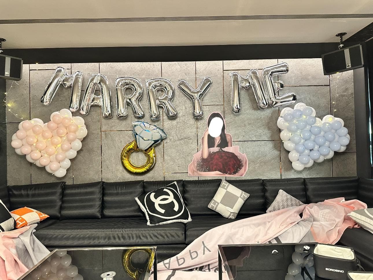 包廂求婚佈置,牆上銀色 MARRY ME 字母,兩側是粉色與藍色愛心氣球,中間是戒指與人形立牌