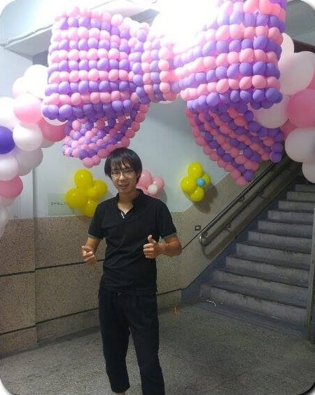 一位穿黑衣的男士站在編織蝴蝶結氣球拱門下比讚