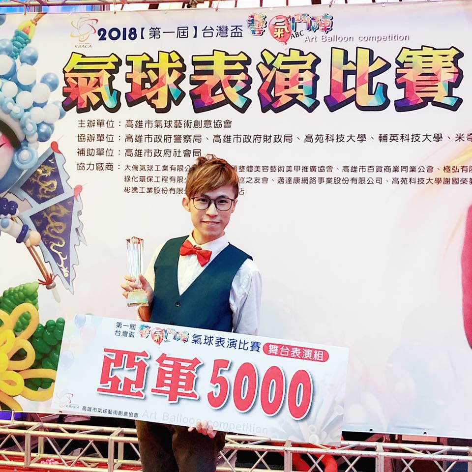 柯男在第一屆台灣盃藝氣鬥陣氣球表演比賽背板前，拿著舞台表演組亞軍獎盃與獎牌板