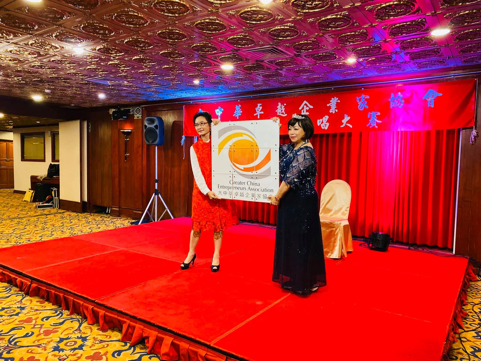 紅色布幕舞台上，兩位女士一起展示大中華卓越企業家協會 Logo 看板