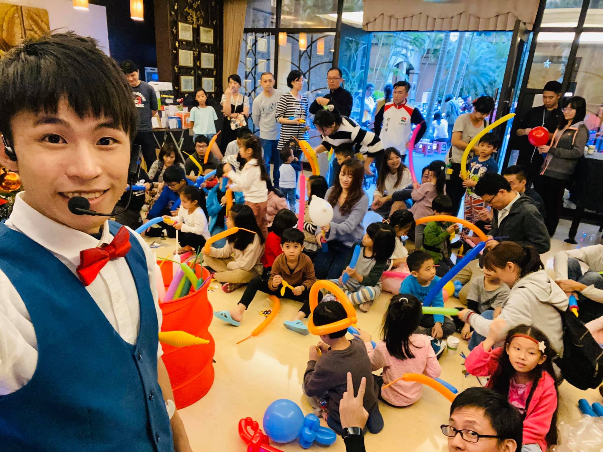 我戴著耳麥自拍，後方孩子和家長坐在地上拿長條氣球做 DIY