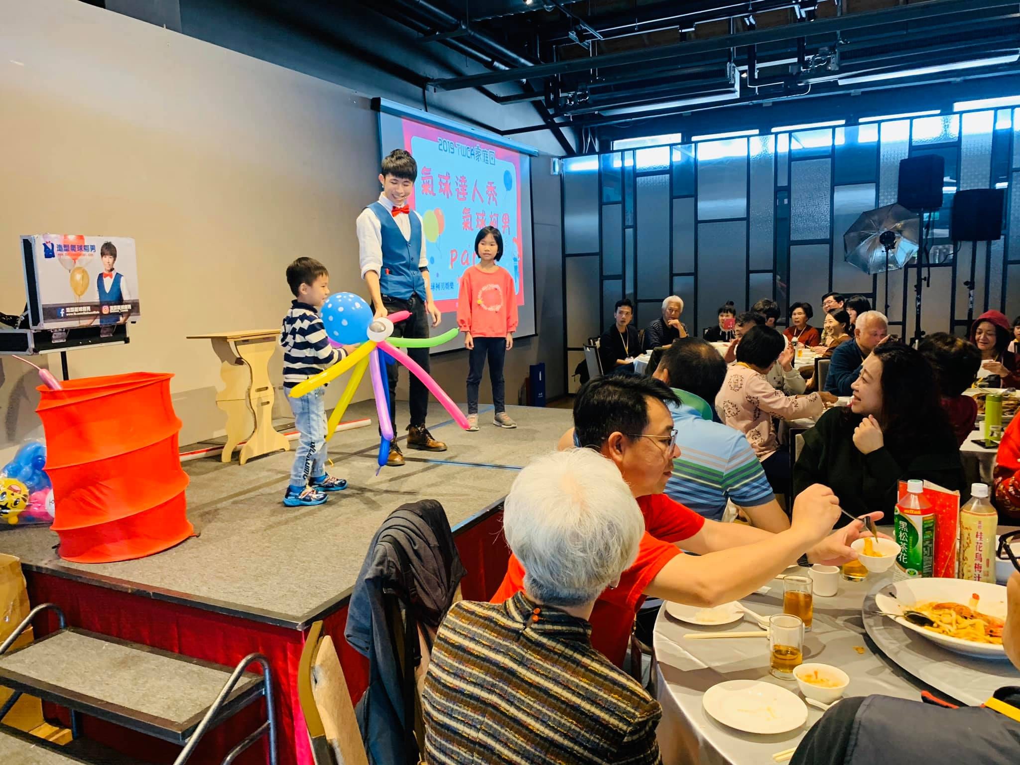餐會舞台上，小男孩站在彩色長條氣球造型旁，台下員工家屬用餐觀看