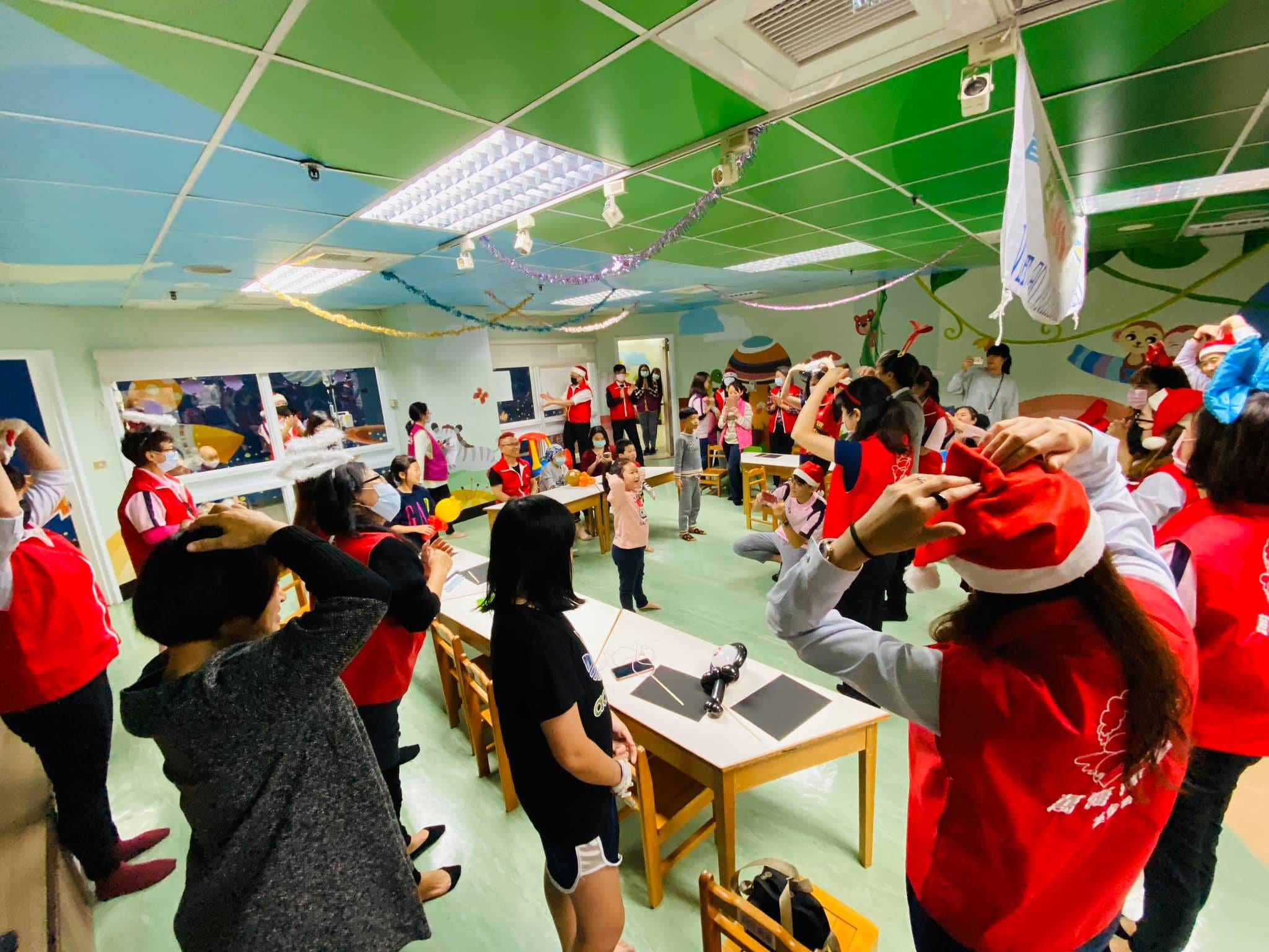 病房活動室裡，戴聖誕帽、穿紅背心的志工和孩子一起做雙手抱頭的互動