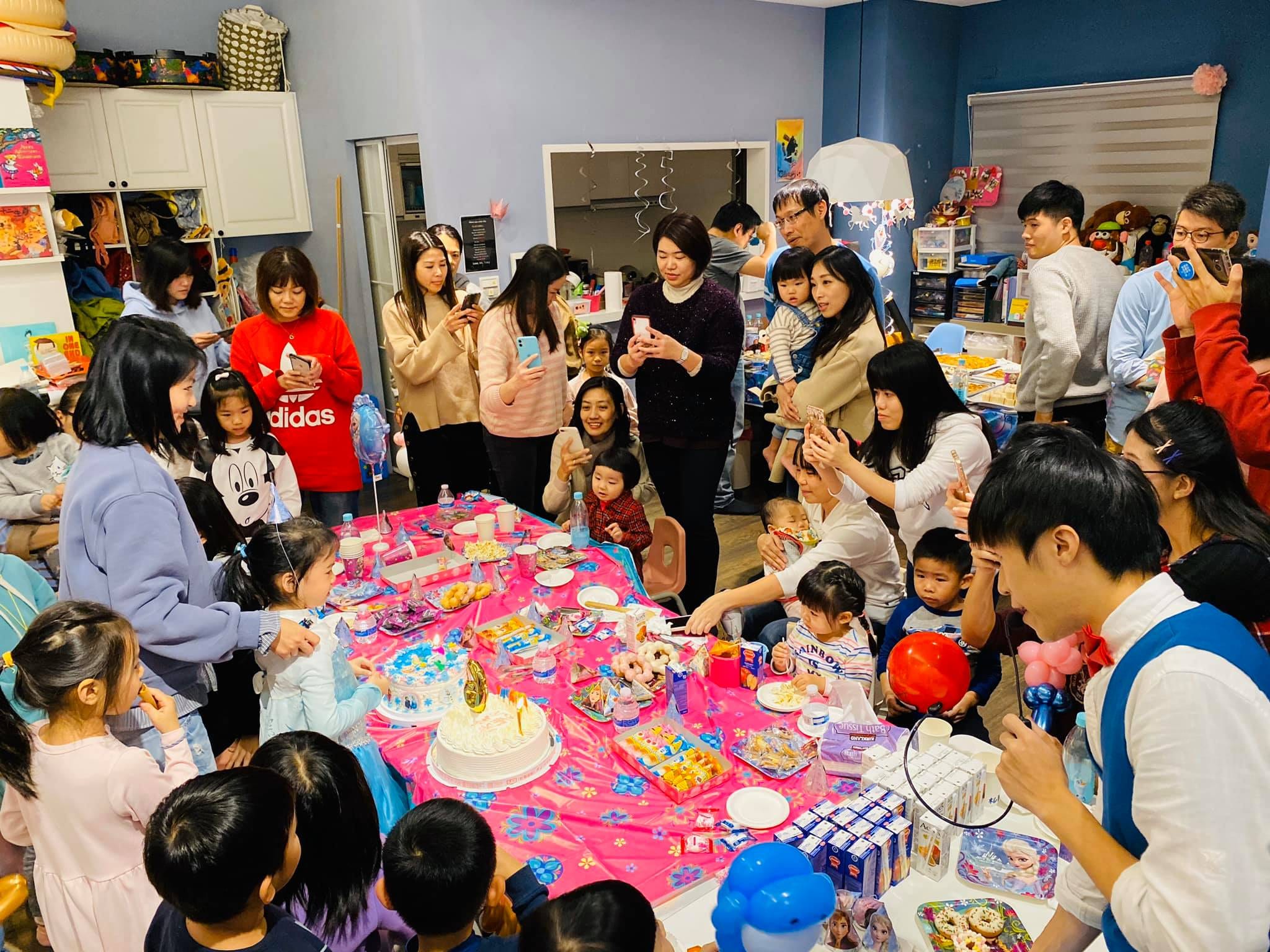 桃紅色桌巾的蛋糕桌旁，大人小孩圍在一起準備唱生日快樂歌