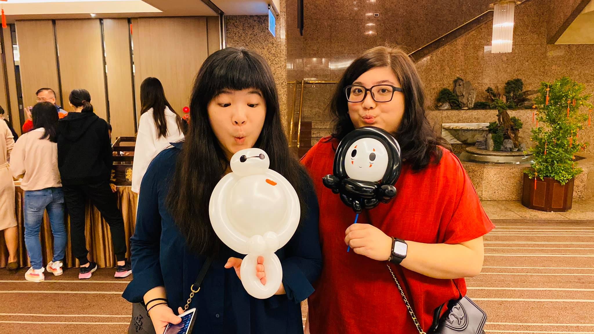 兩位女生在宴會廳外，分別拿著白色杯麵與黑白配色的圓臉氣球
