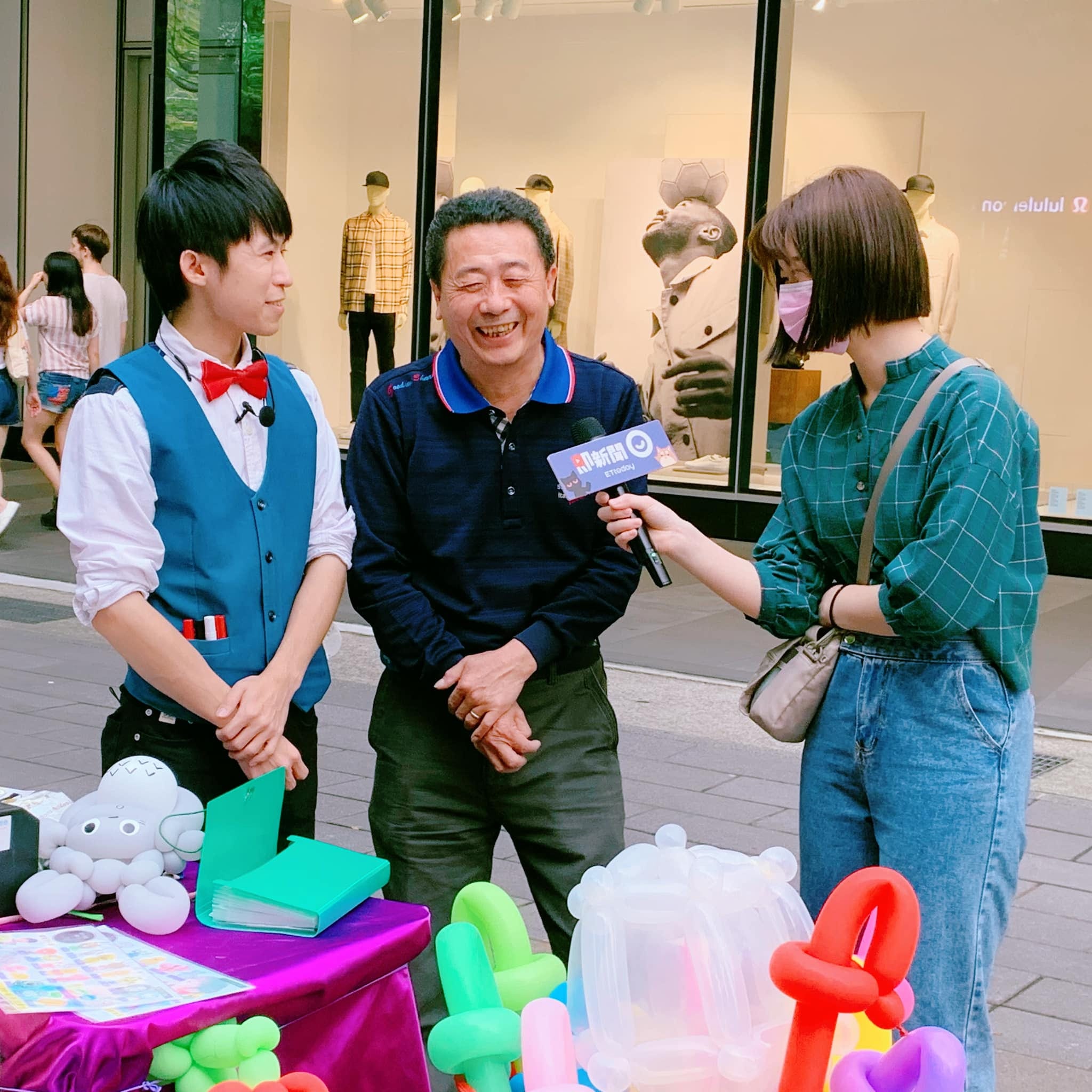 即新聞記者拿麥克風採訪我和爸爸柯爹，攤位桌上擺著造型氣球